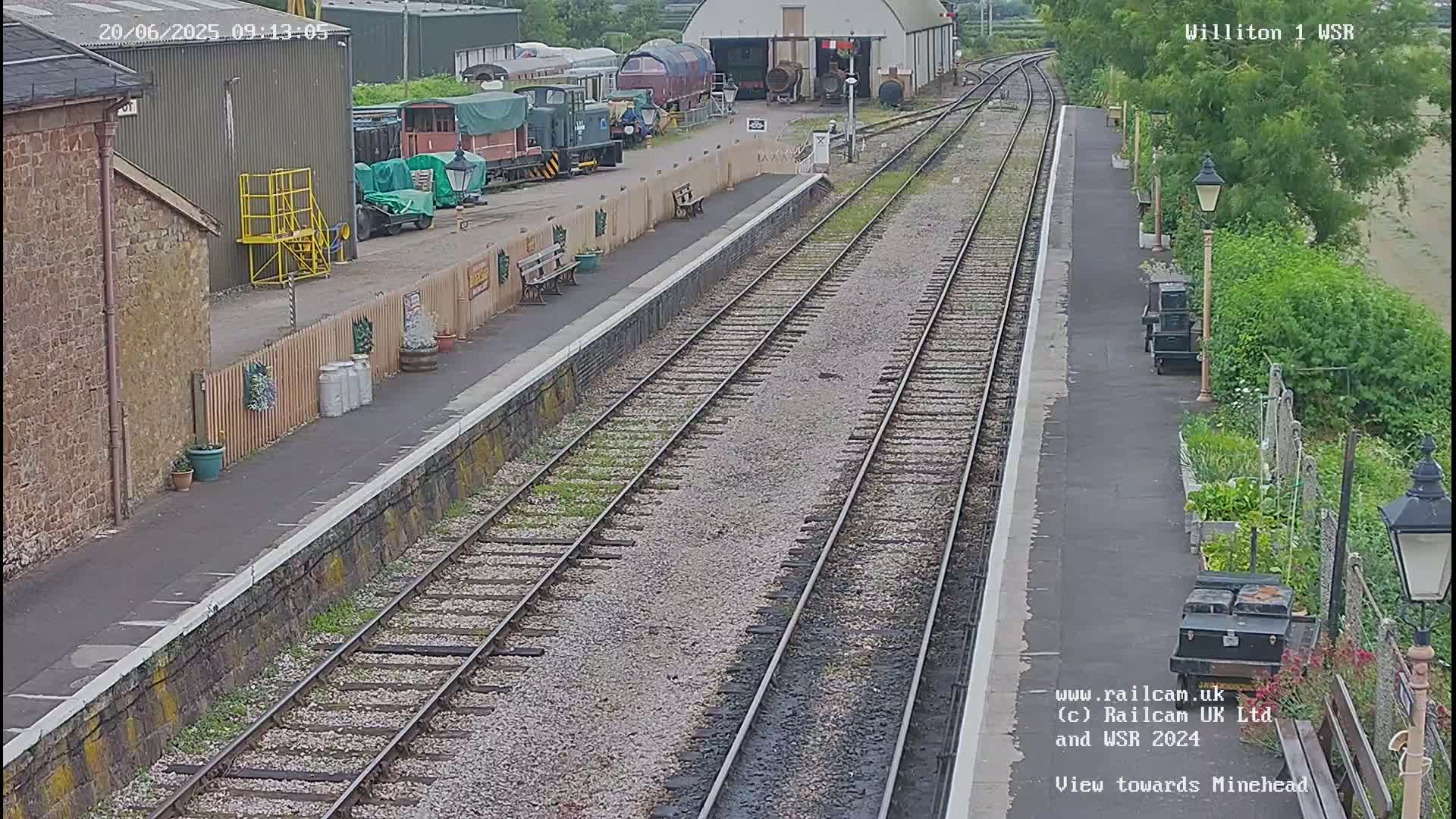 A railway station platform with multiple train tracks stretches into the distance on a partly sunny day.