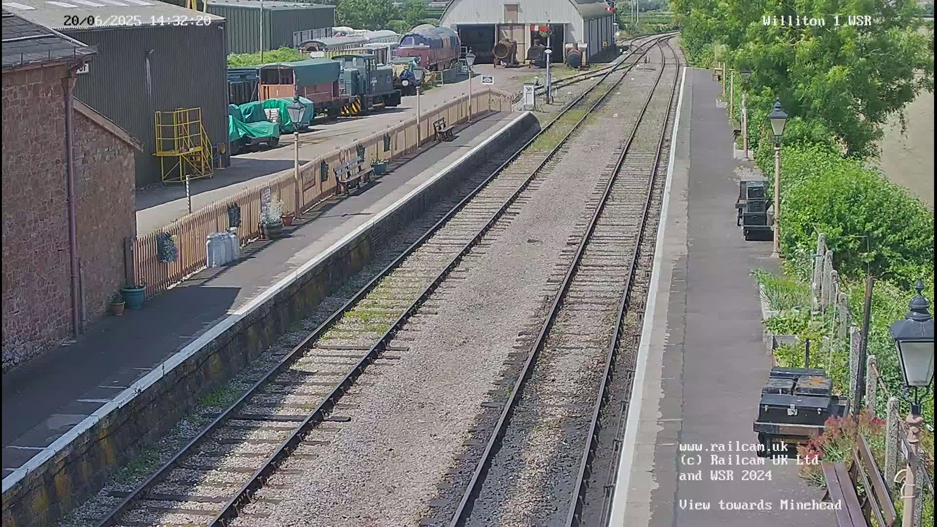 A sunny day at a train station shows railway tracks extending into the distance, with several train cars parked near a building.