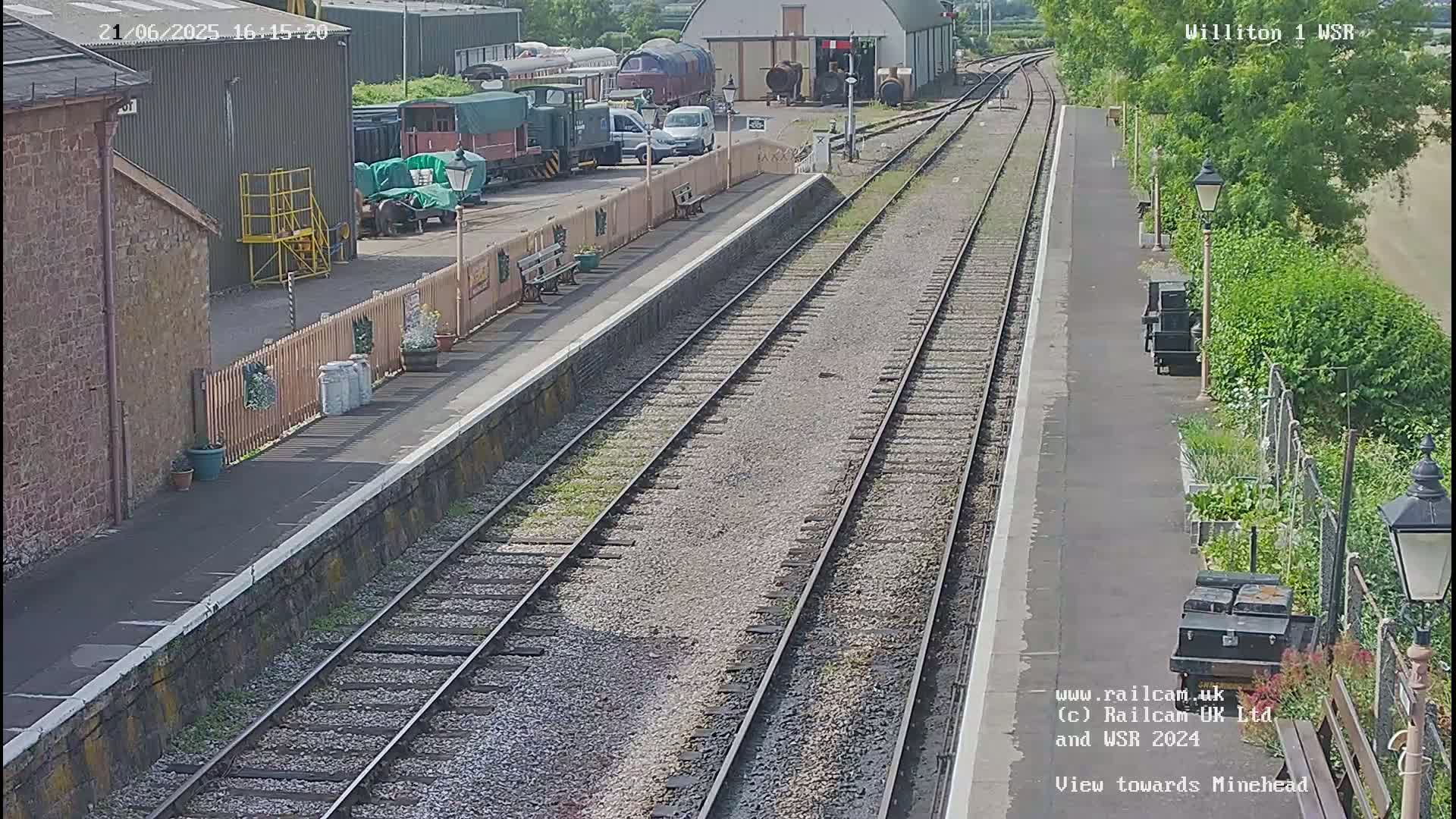 A sunny view of a train station platform with multiple train cars and some vehicles parked nearby, along with a small garden area.