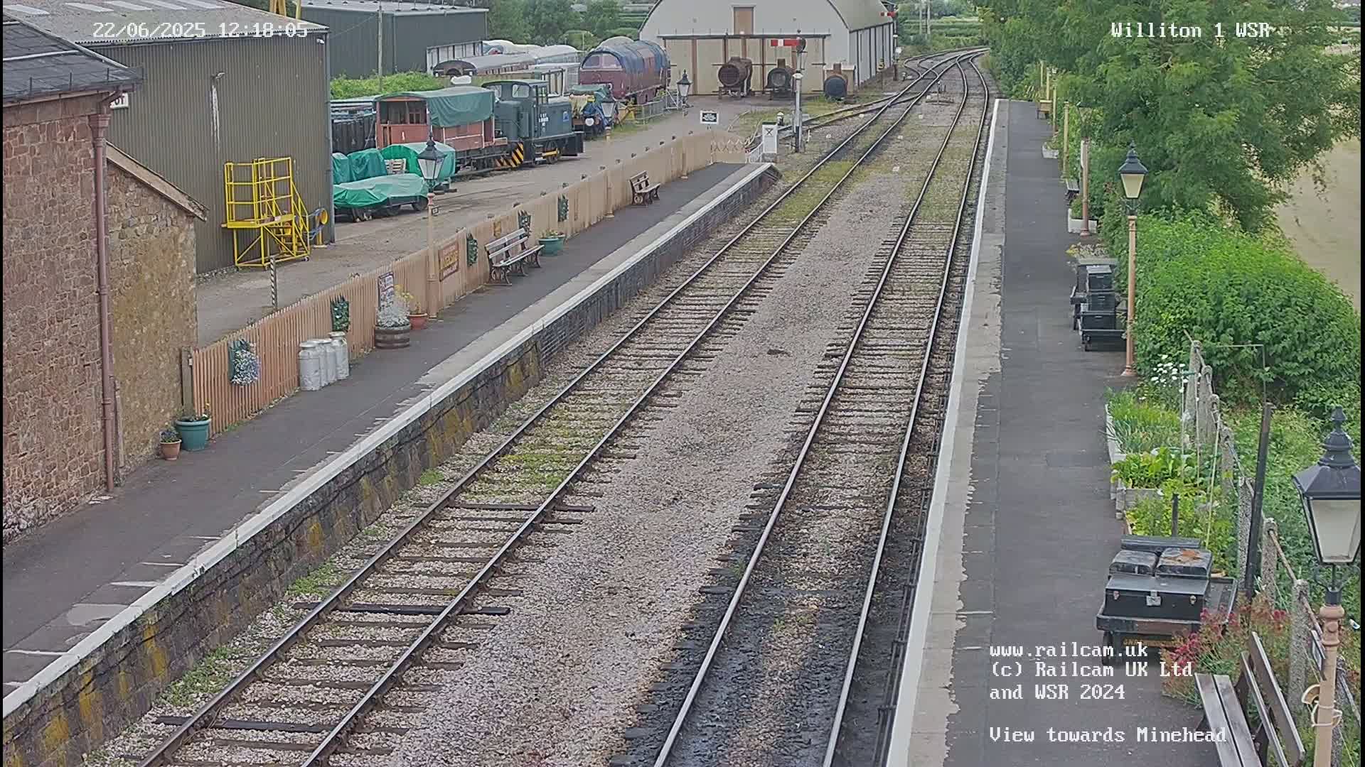 A railway station platform with multiple train tracks extends into the distance on an overcast day, featuring several parked trains and a small garden beside it.