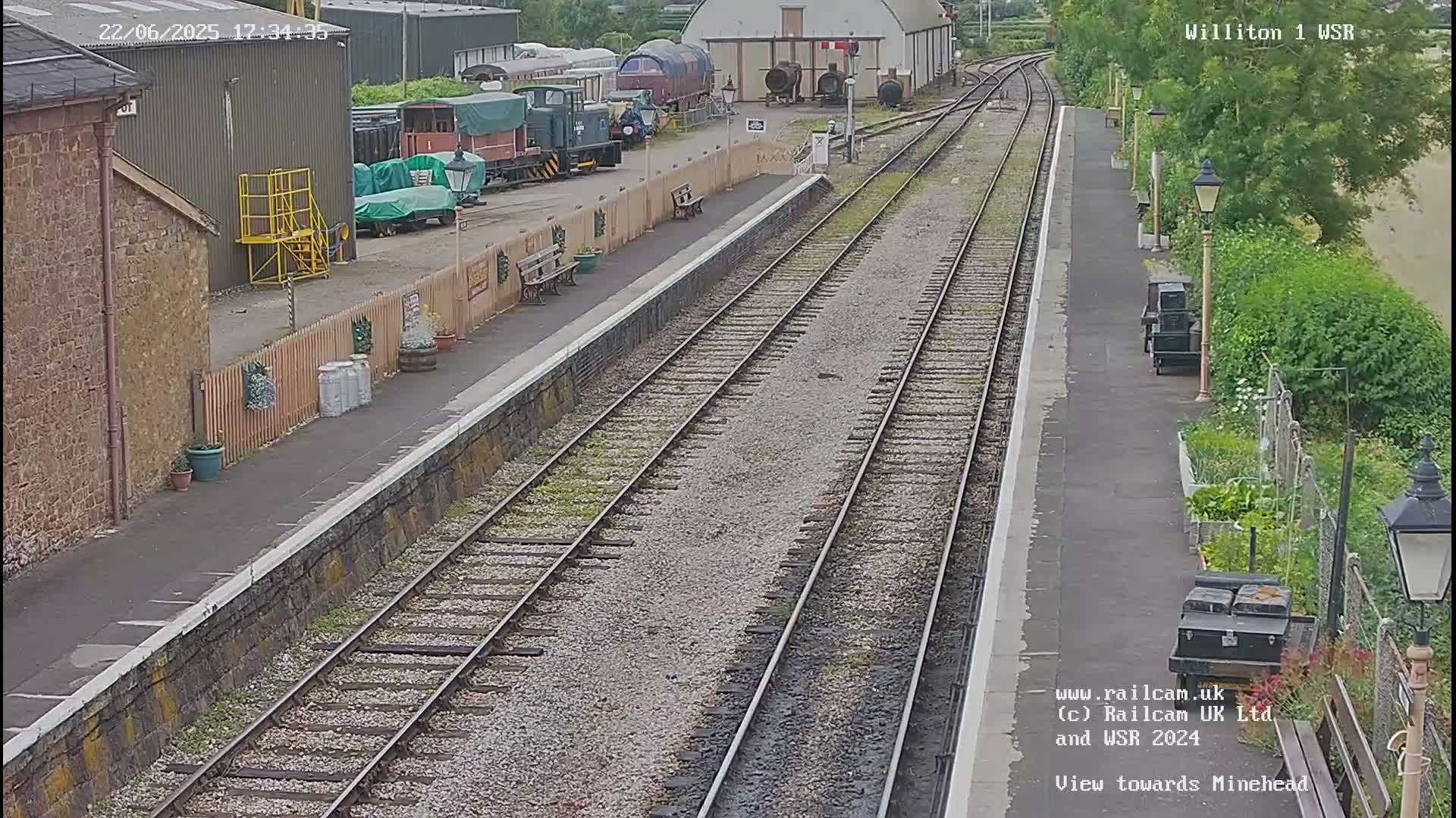 A train station platform with multiple train tracks and railway cars visible, under a partly sunny sky.
