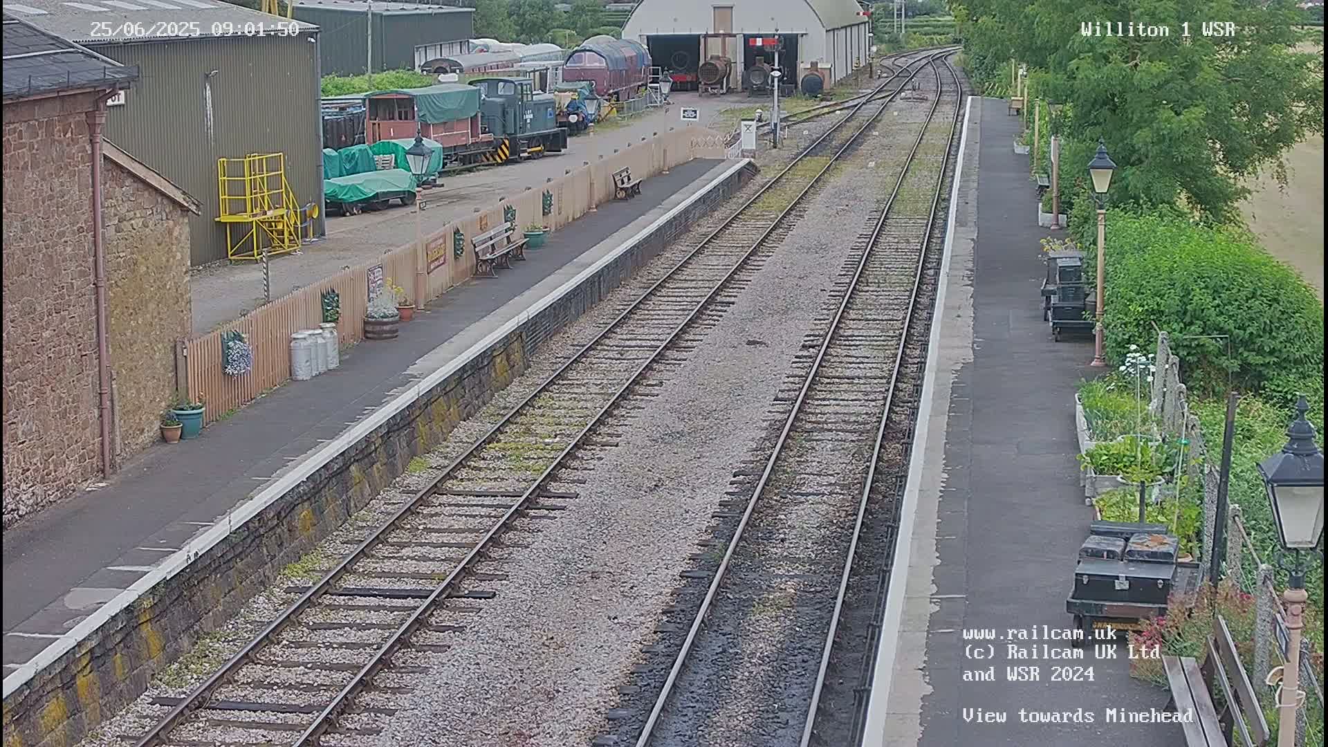 A train station platform with multiple railway tracks extends into the distance on a partly cloudy day, with various railway cars and buildings visible nearby.