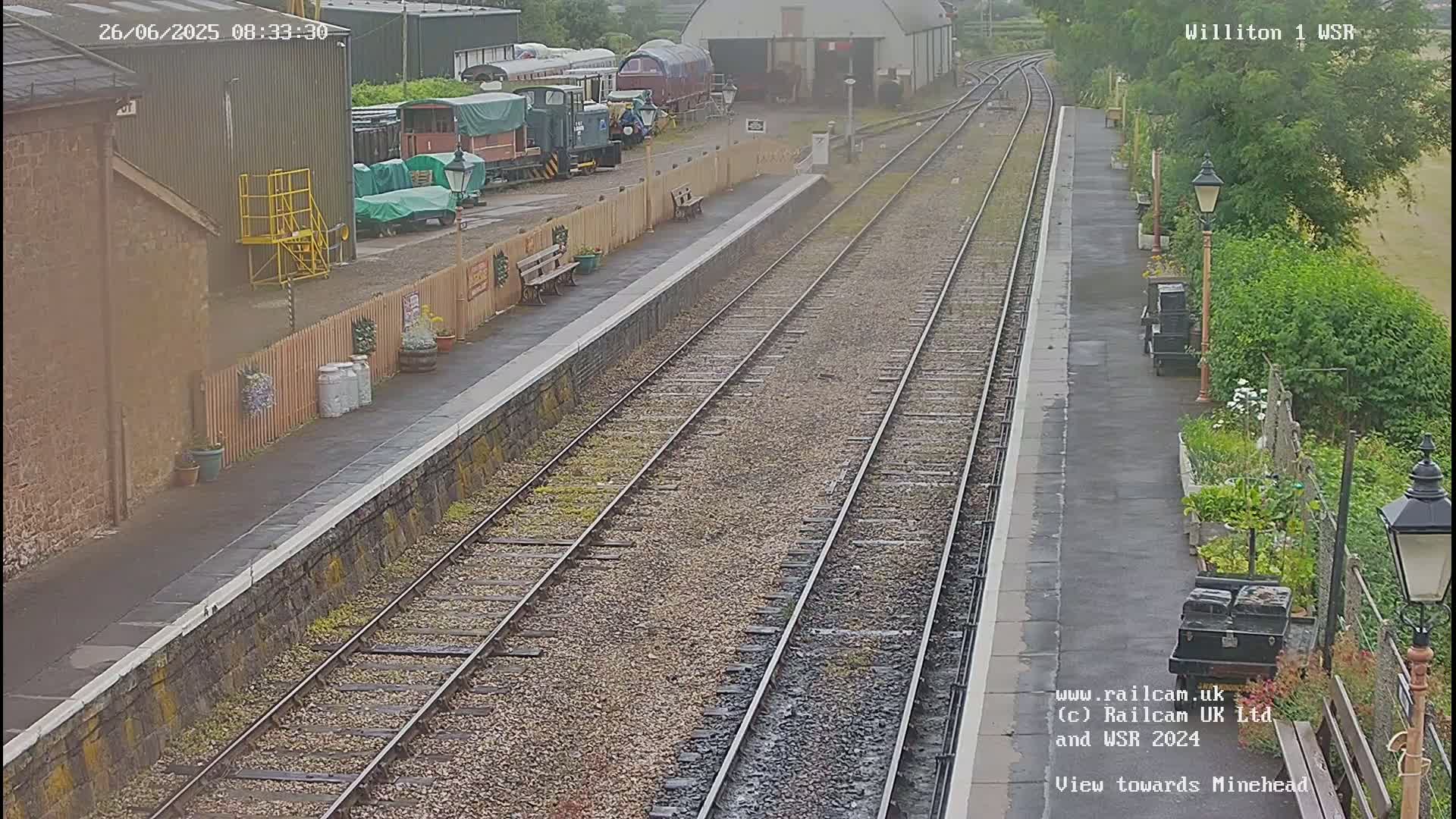 A train station platform with multiple train tracks, railway cars, and a small garden is seen on an overcast day.