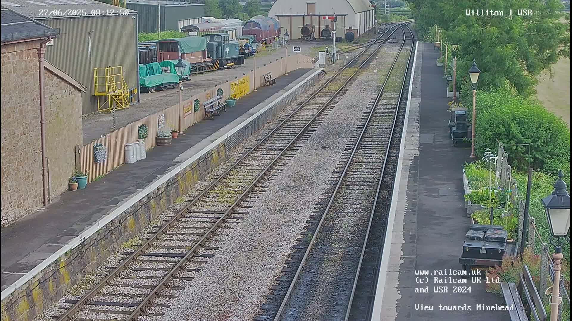 A train station platform with multiple train tracks and several railway carriages visible in the background on a sunny day.