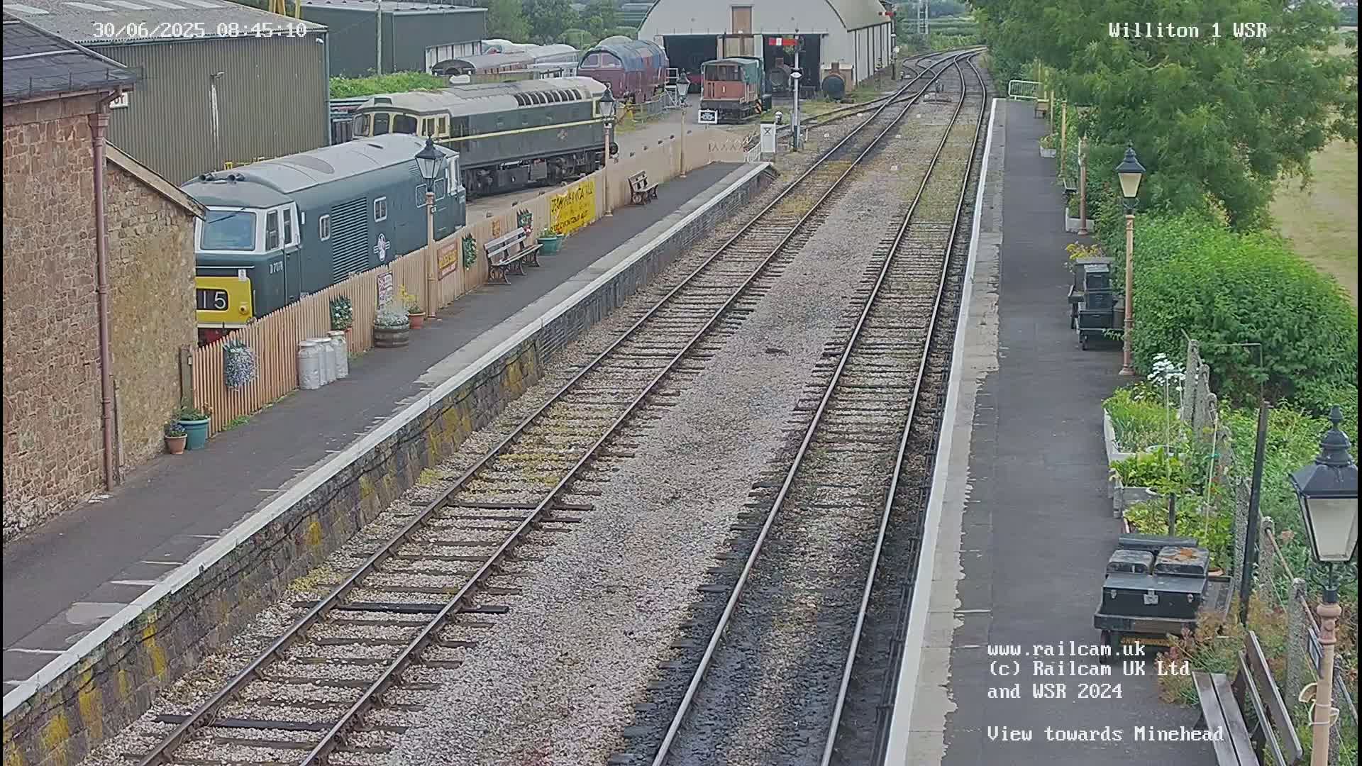 A train station on a partly sunny day with several train cars parked on the tracks, a train partially visible behind a fence, and a garden next to the platform.