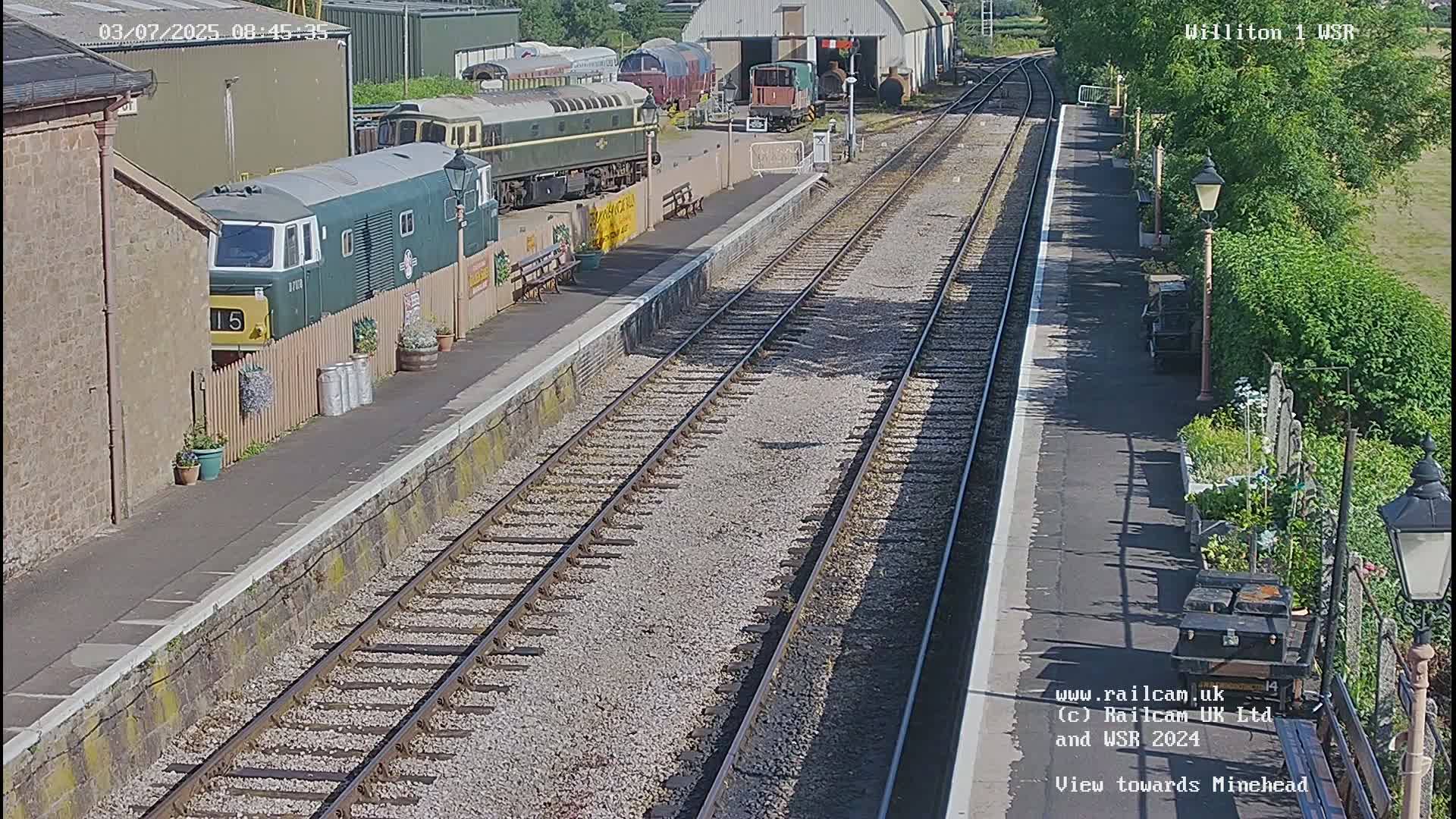 A sunny day at a train station with multiple train cars parked near a platform and railway tracks extending into the distance.