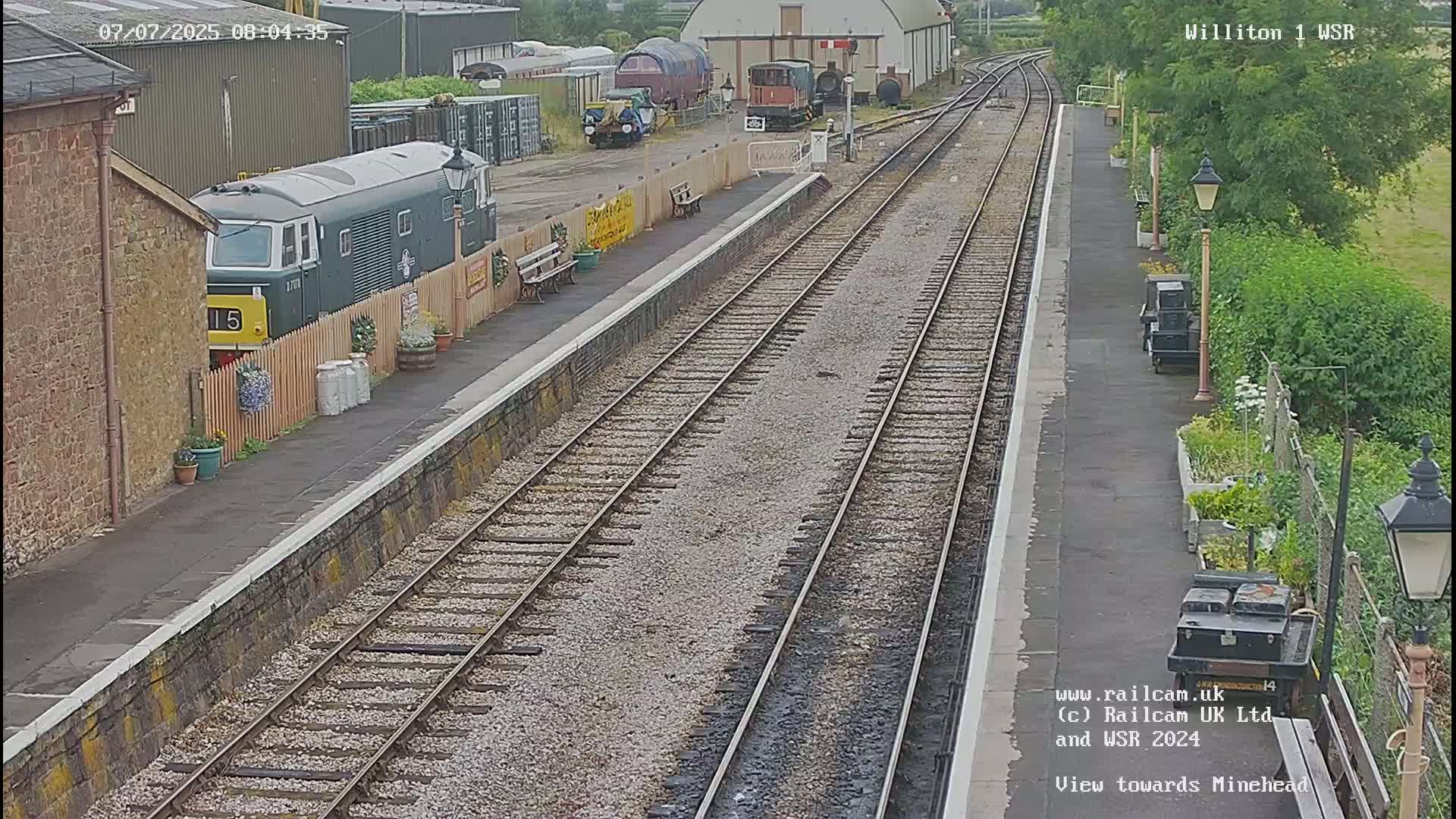 A train station platform with a diesel locomotive parked beside it, multiple train tracks extending into the distance, and  a partly sunny day.