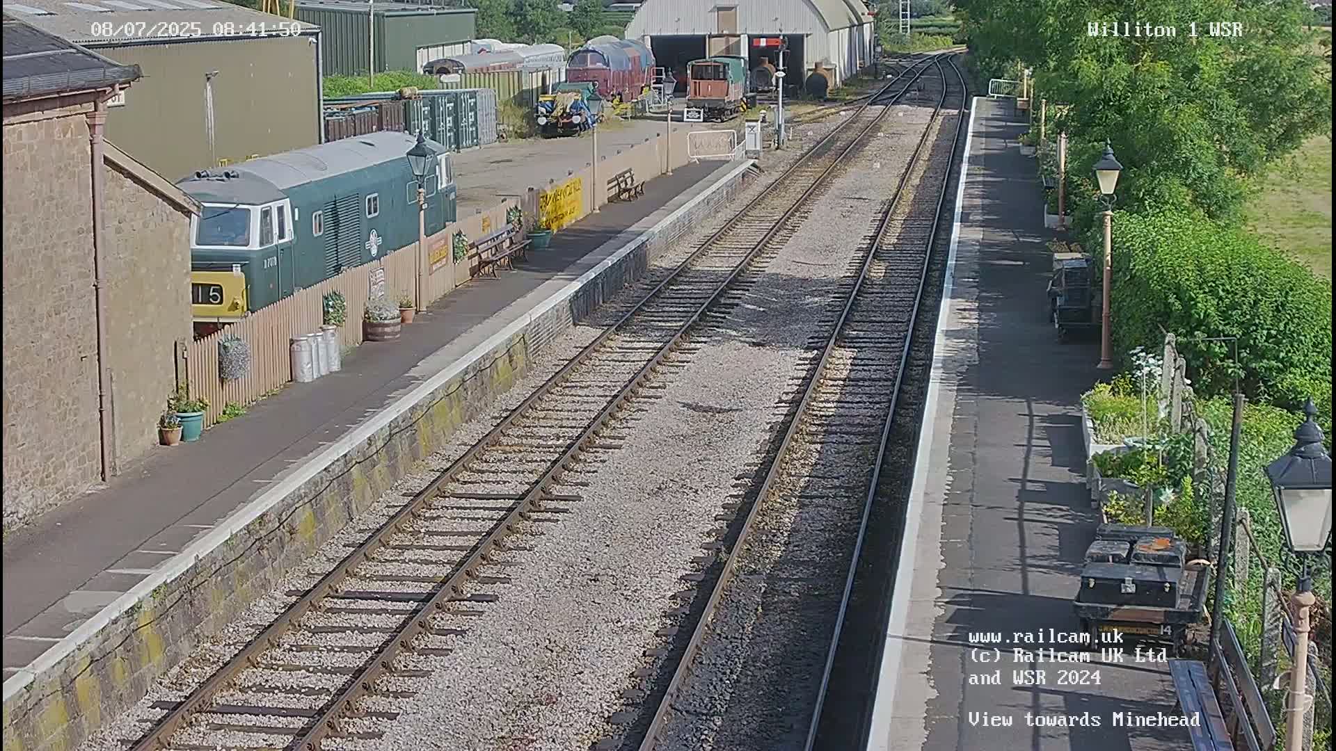 A sunny day at a train station shows a green diesel locomotive parked next to a platform with railway tracks extending into the distance.