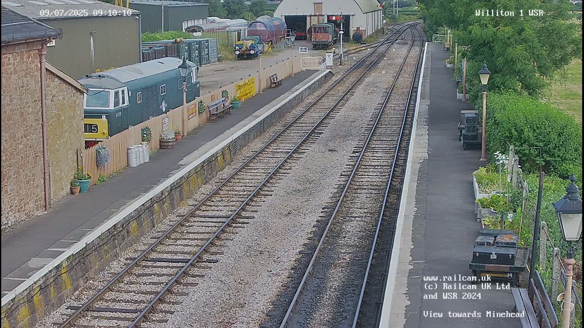 A train station platform with multiple train tracks, a diesel locomotive partially visible behind a fence, and a garden along the side on a partly sunny day.