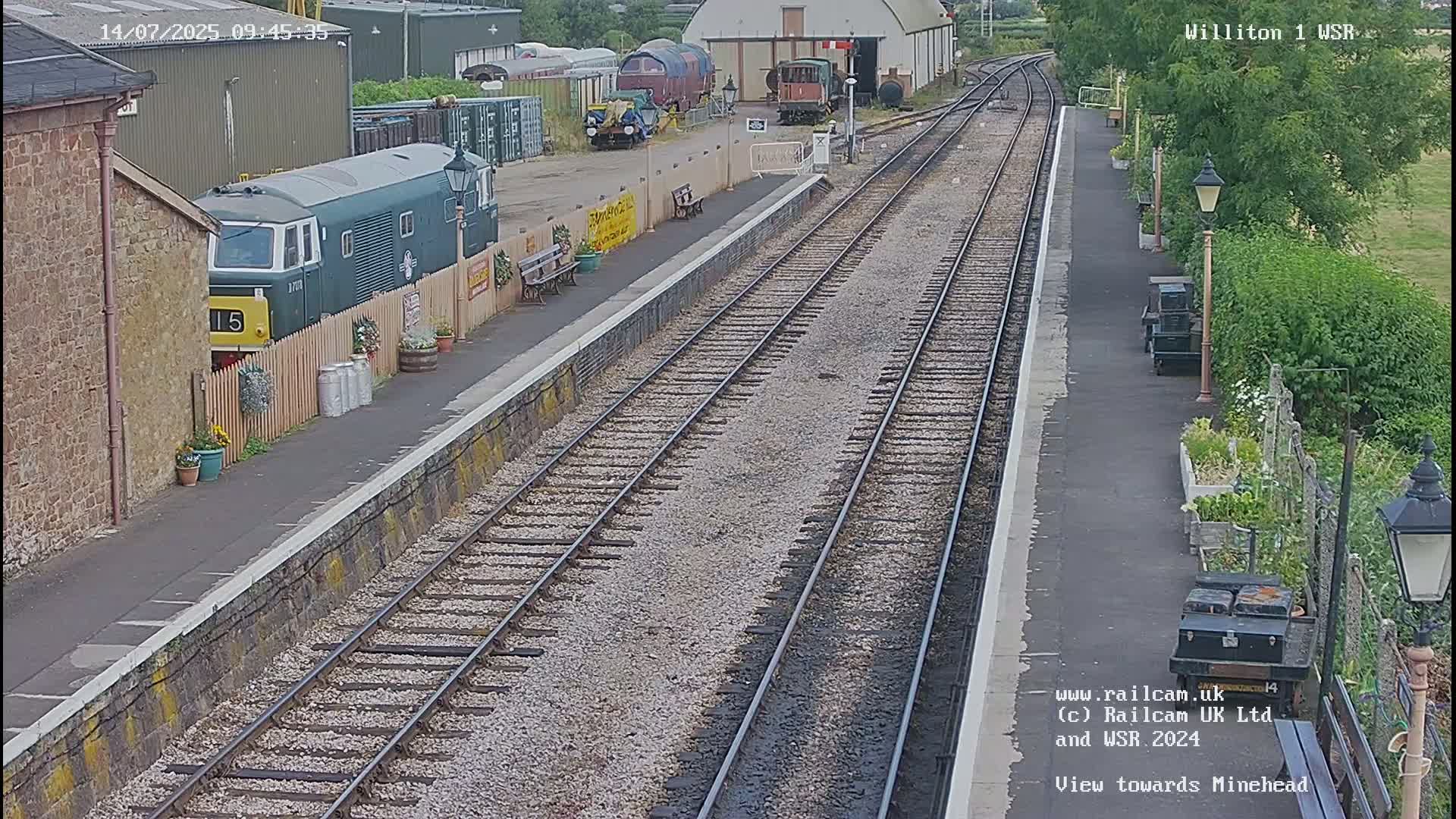 A green diesel locomotive sits at a train station platform next to railway tracks on a partly sunny day.