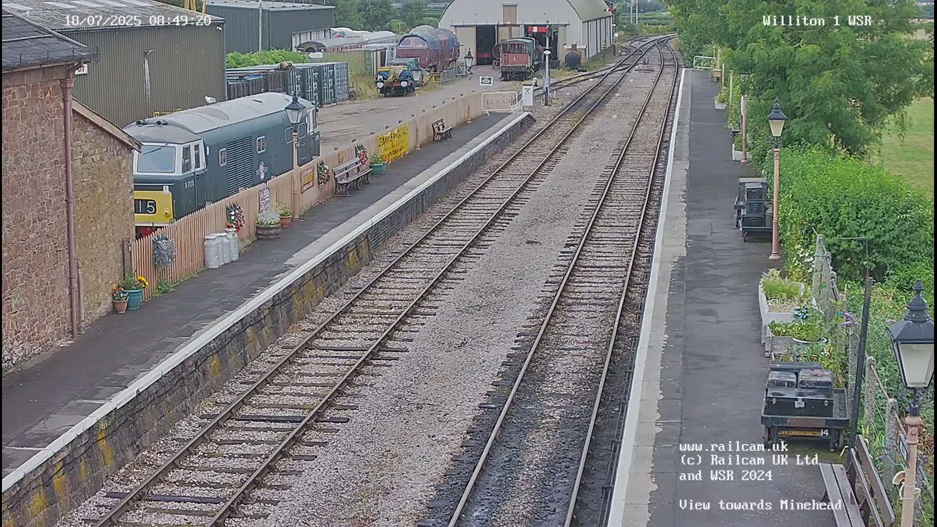 A green diesel locomotive is parked at a train station platform on a partly cloudy day, with multiple railway tracks extending into the distance.