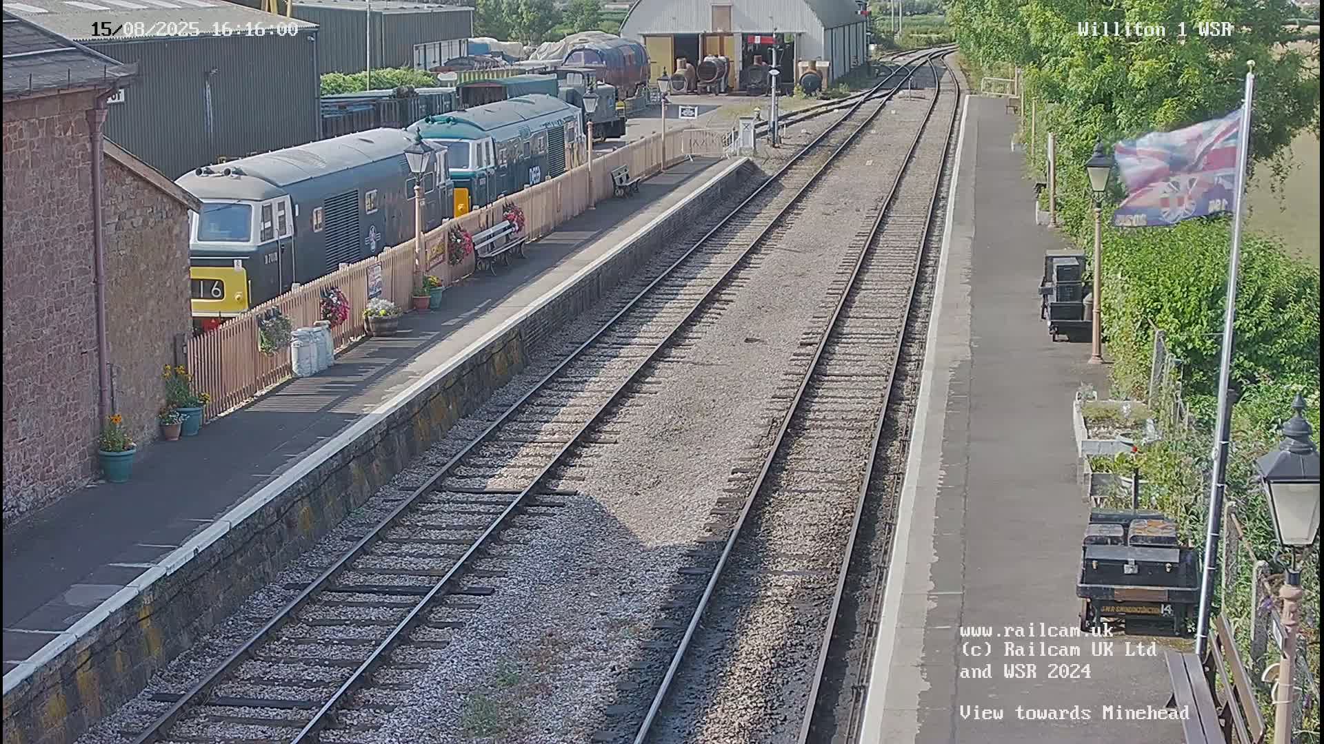A green diesel locomotive is parked at a train station platform on a partly cloudy day, with multiple railway tracks extending into the distance.