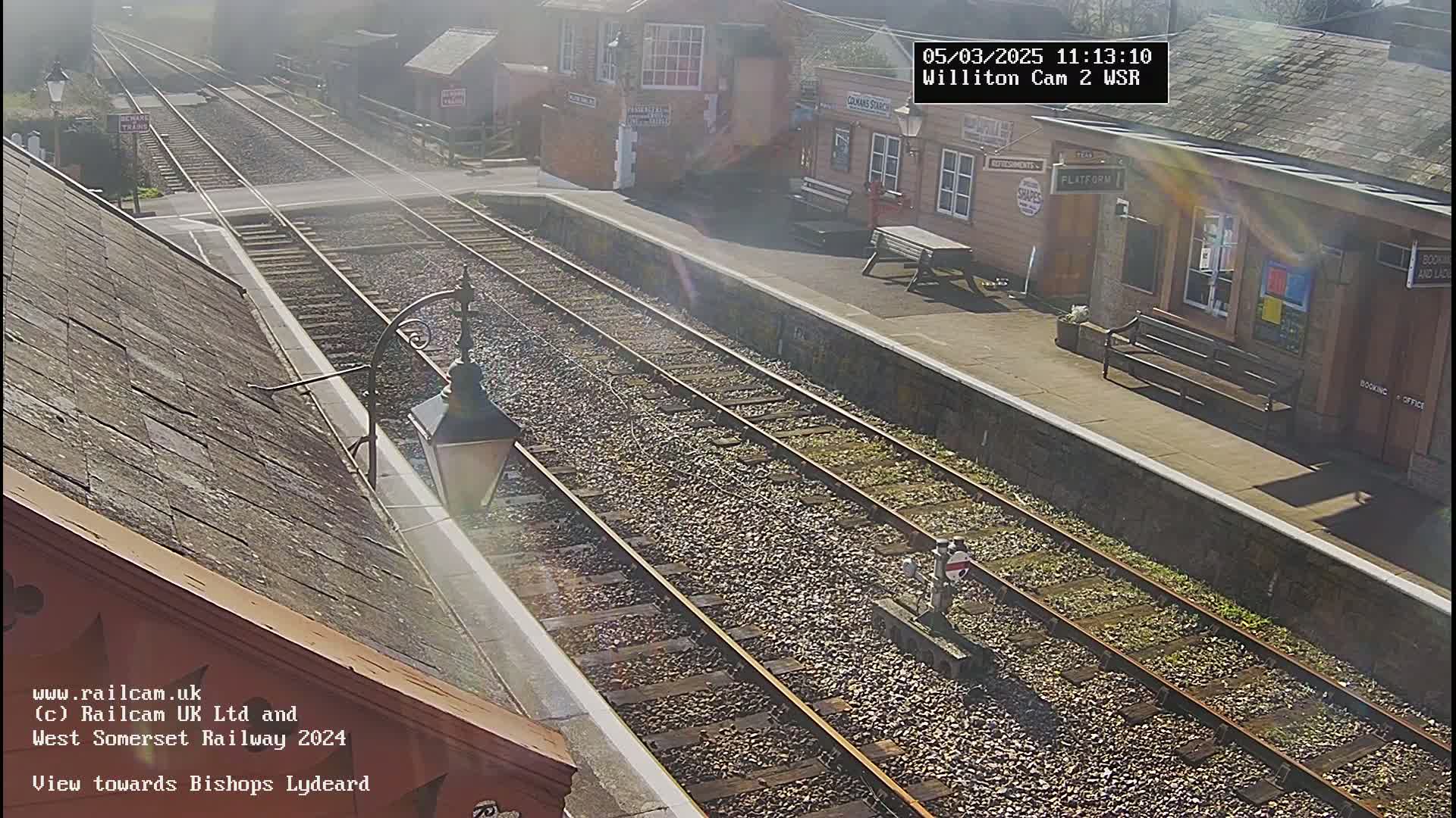 A sunny view of a train station platform with two sets of train tracks extending into the distance.