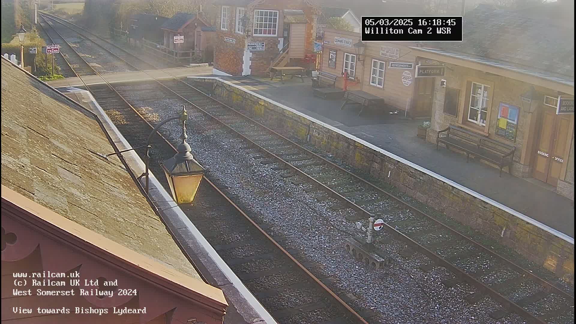 A high-angle view shows a train station on a sunny day with two train tracks extending into the distance.