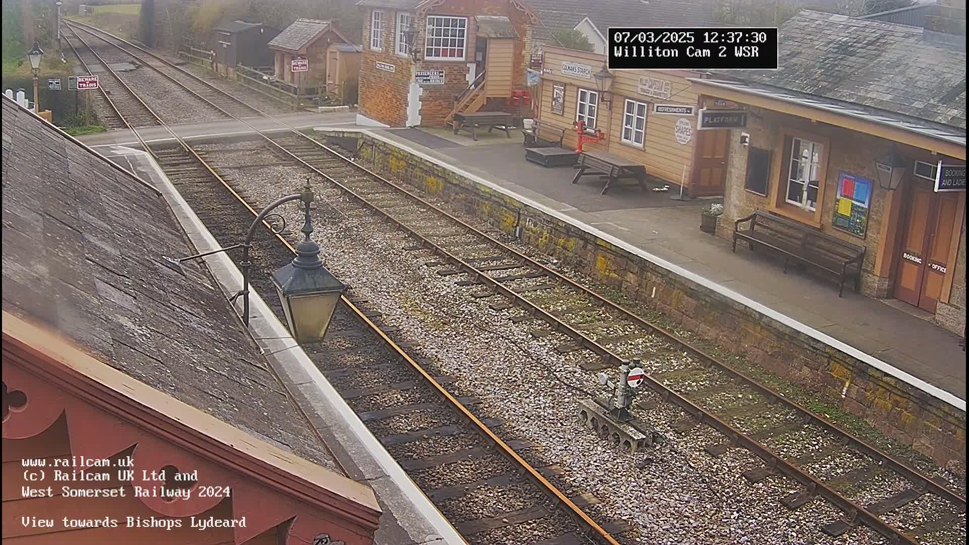 A high-angle view of a train station platform with multiple train tracks, a small building, and overcast weather conditions.