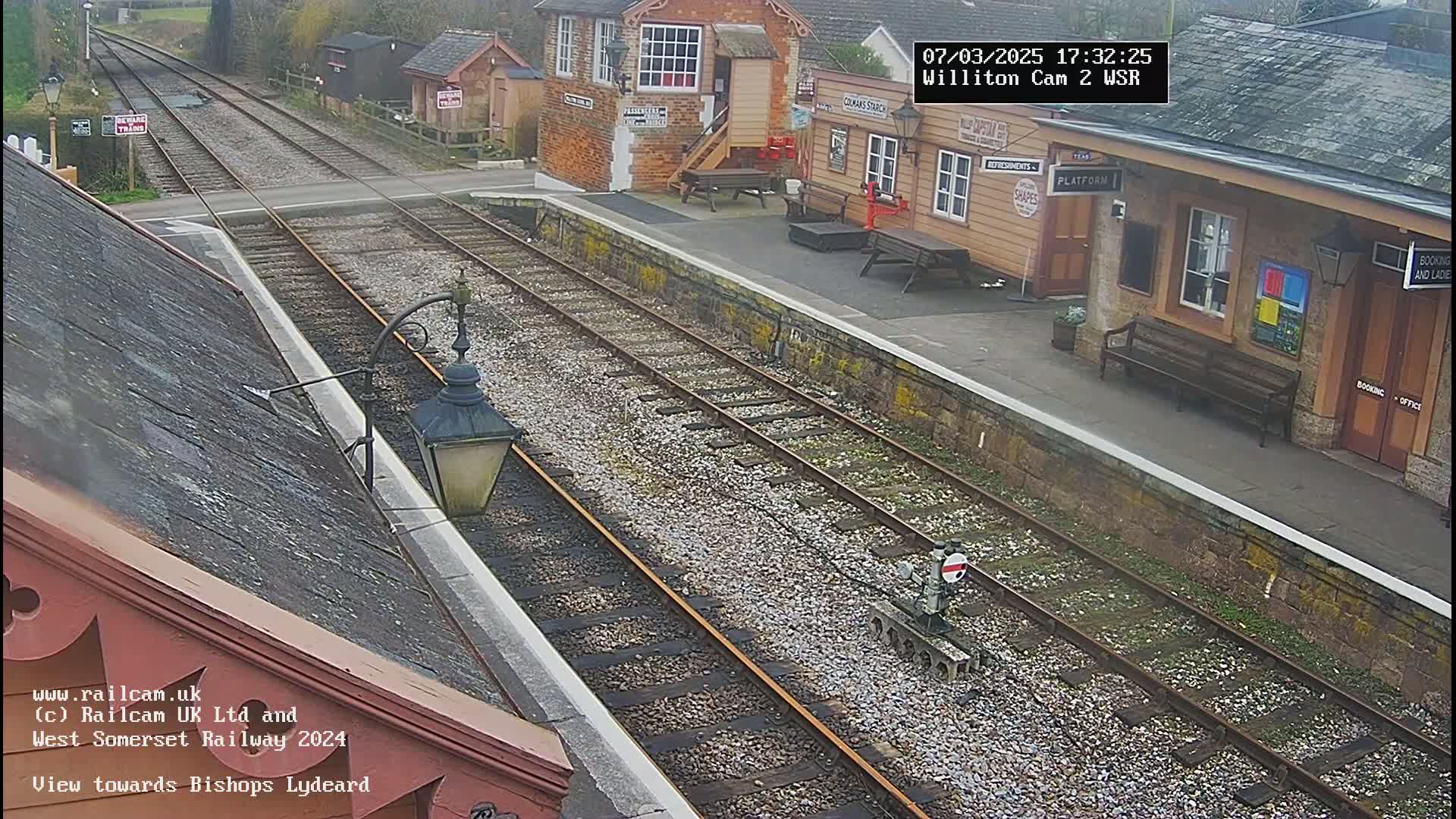 A view of a train station platform and railway tracks on an overcast day, with a small building and a bench visible.