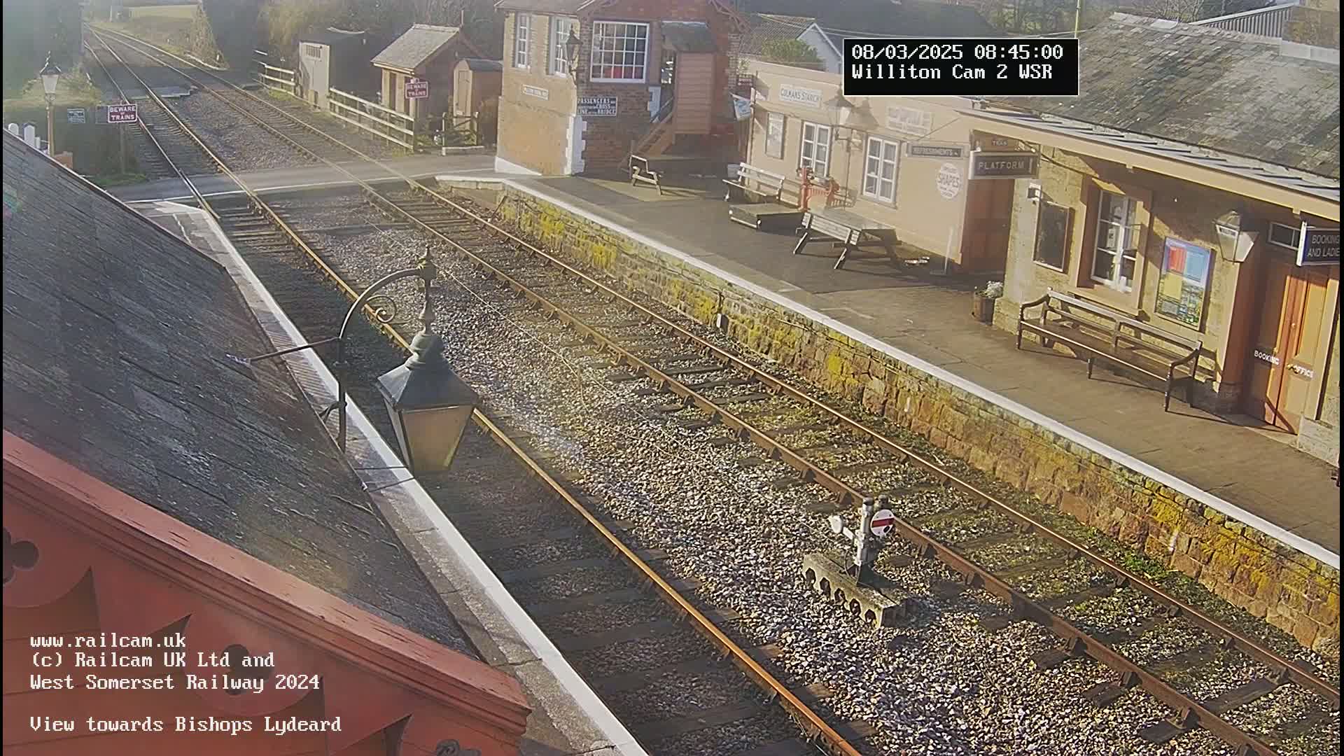 A sunny view of a train station platform with a pair of train tracks extending into the distance.
