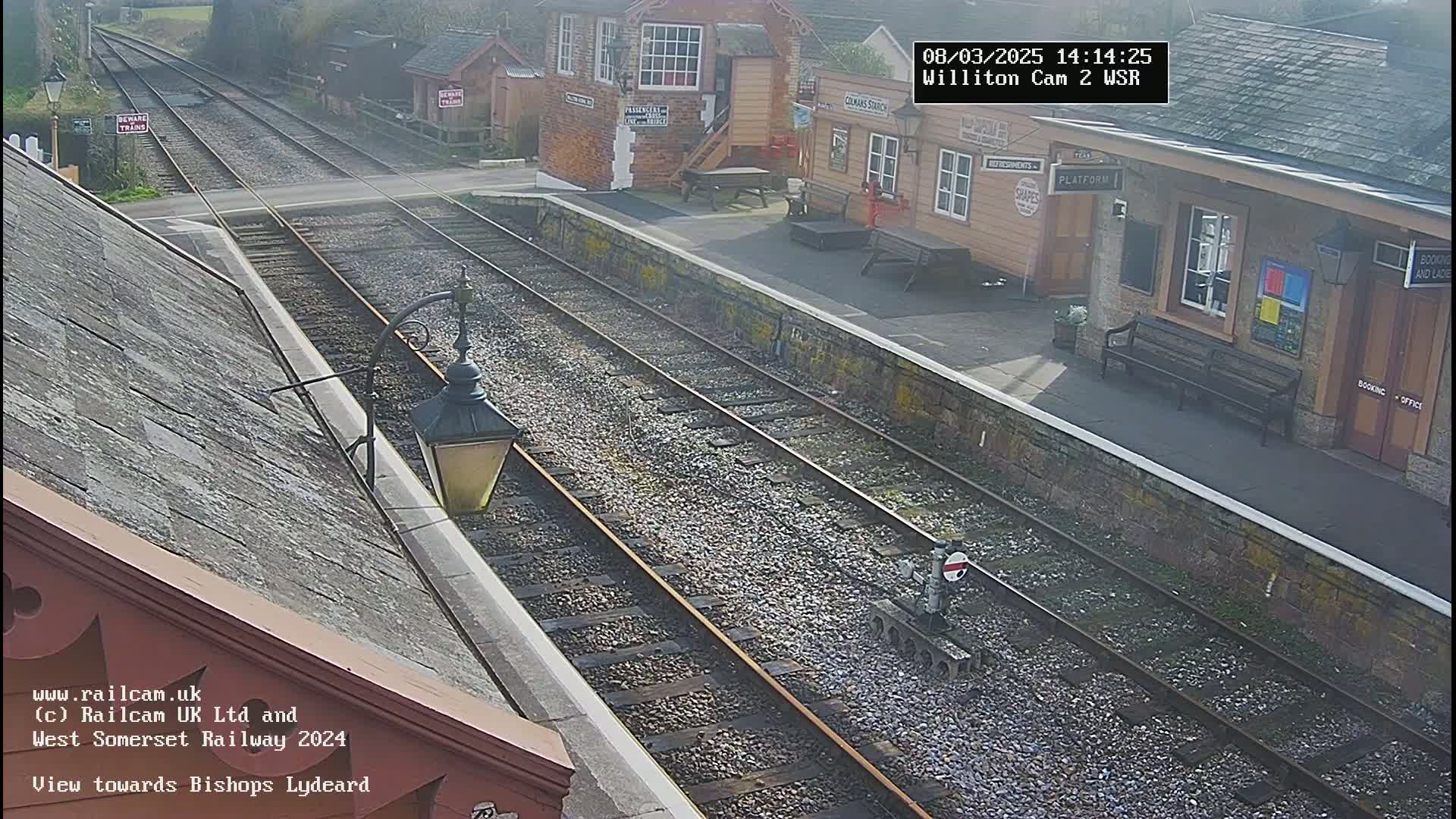 A sunny view of a train station platform and railway tracks, with a building visible in the background.