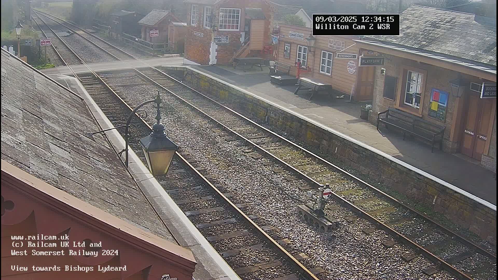 A high-angle view of a train station on a sunny day shows two parallel train tracks leading away from the station, which has a small building and a bench.