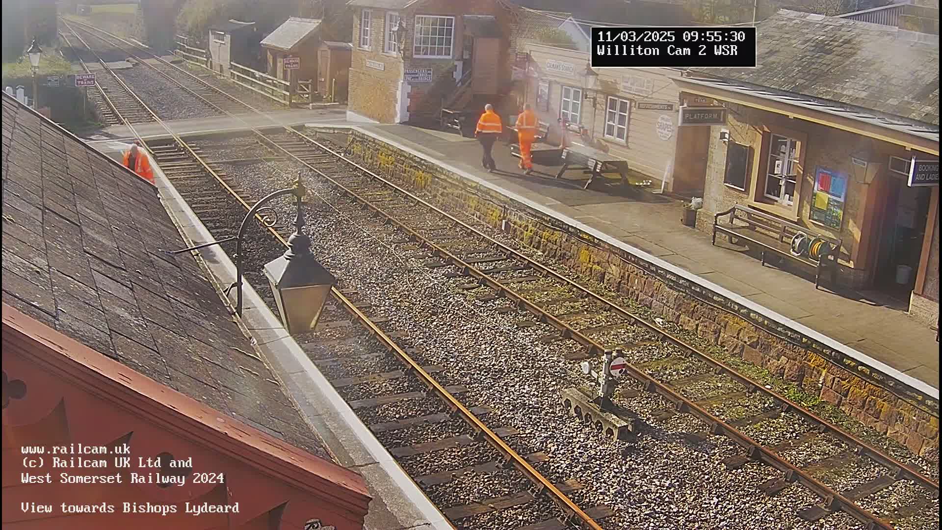 A sunny view of a train station platform with two workers in orange vests and a section of train tracks extending into the distance.