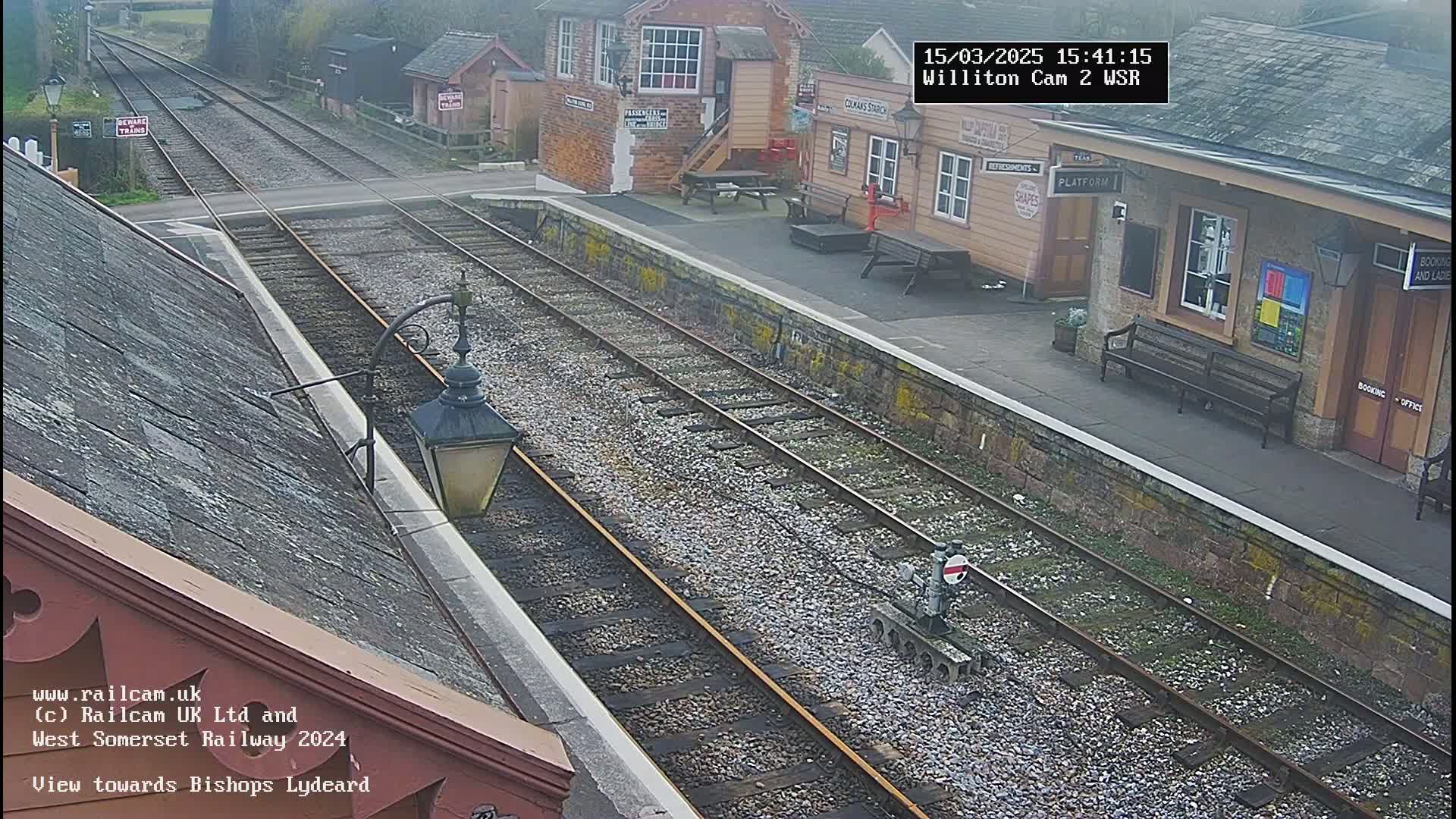 A high-angle view of a train station platform on an overcast day, showing train tracks extending into the distance.