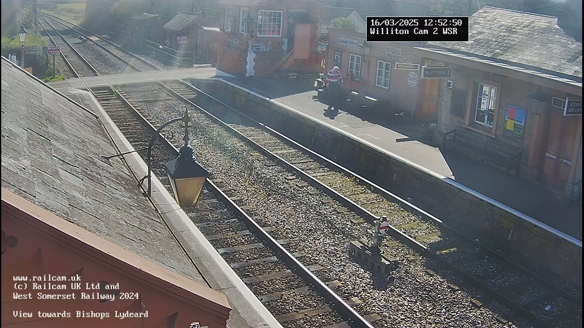 A sunny view of a train station platform with railroad tracks, a building, and a person standing on the platform.