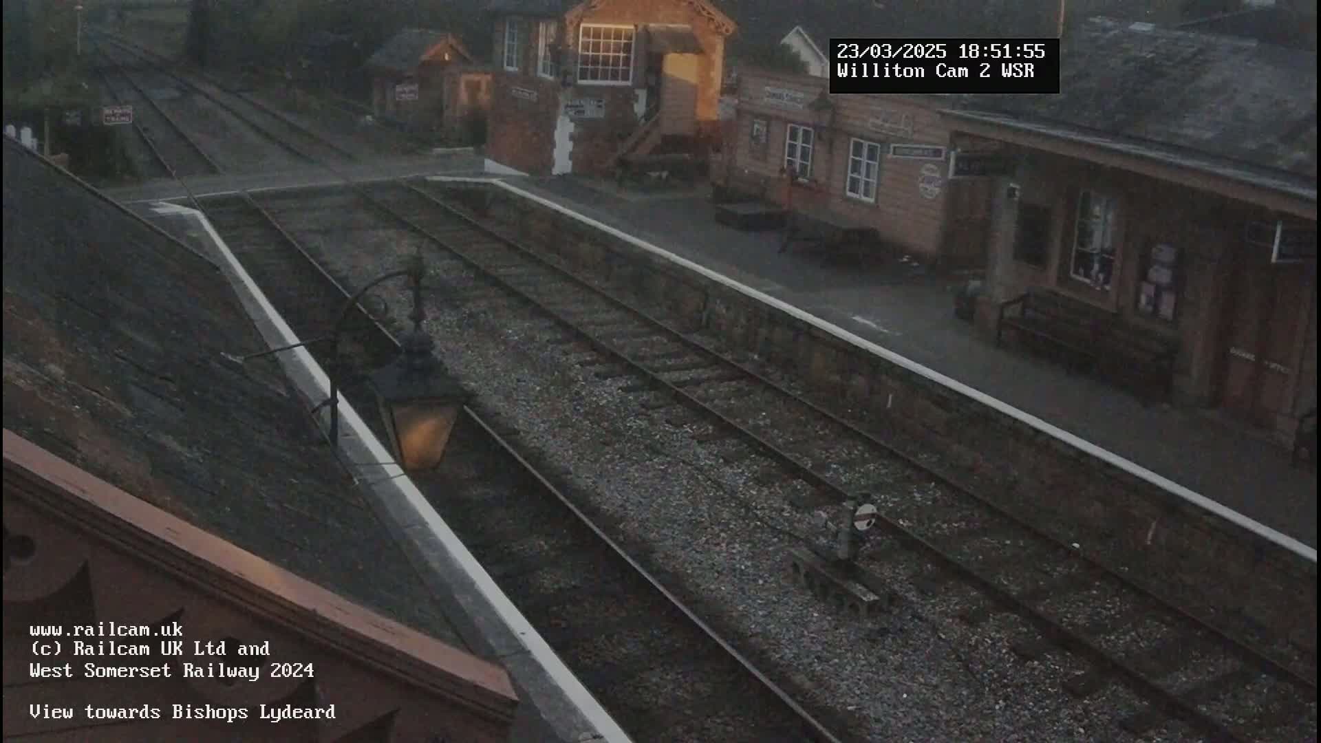 A nighttime view of a train station platform with train tracks and a building under overcast skies.