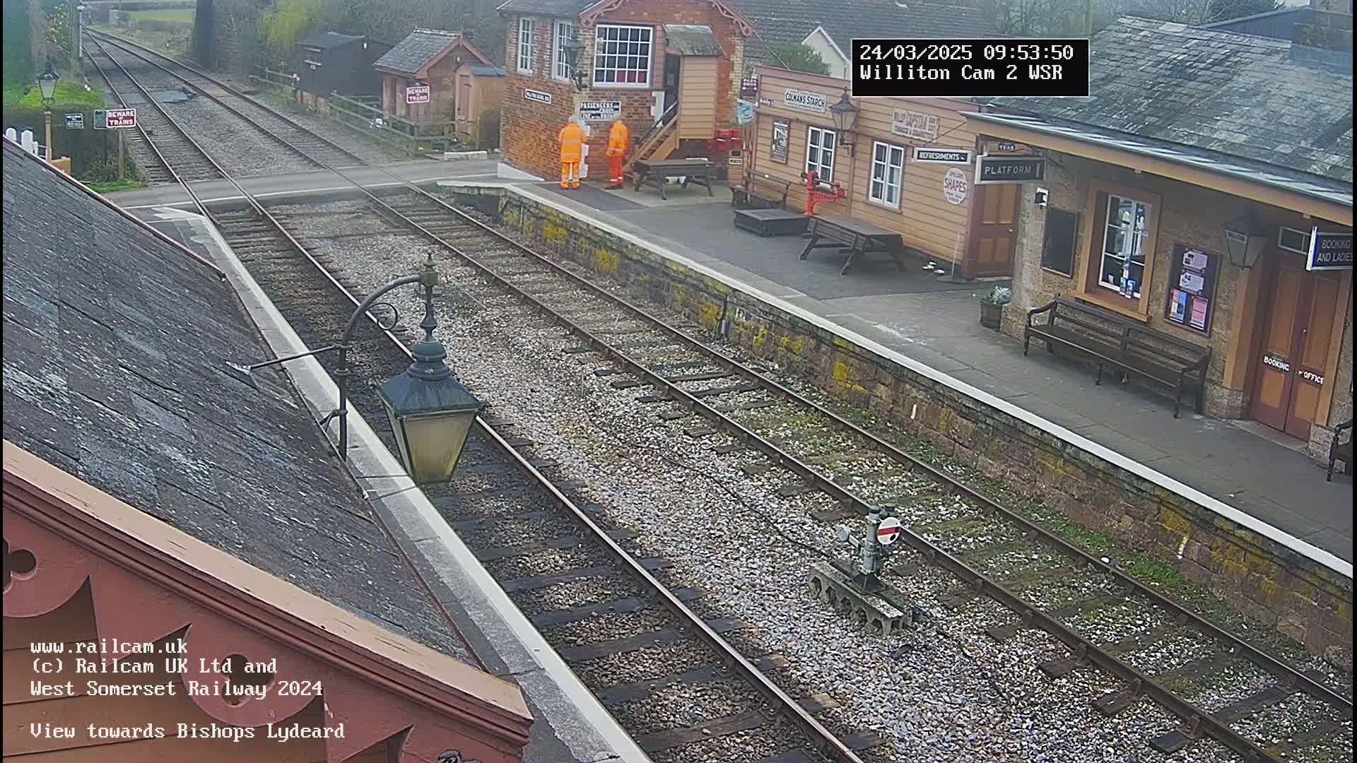 A train station platform with two sets of train tracks, a small railcar, and two people in orange vests is viewed from above on an overcast day.