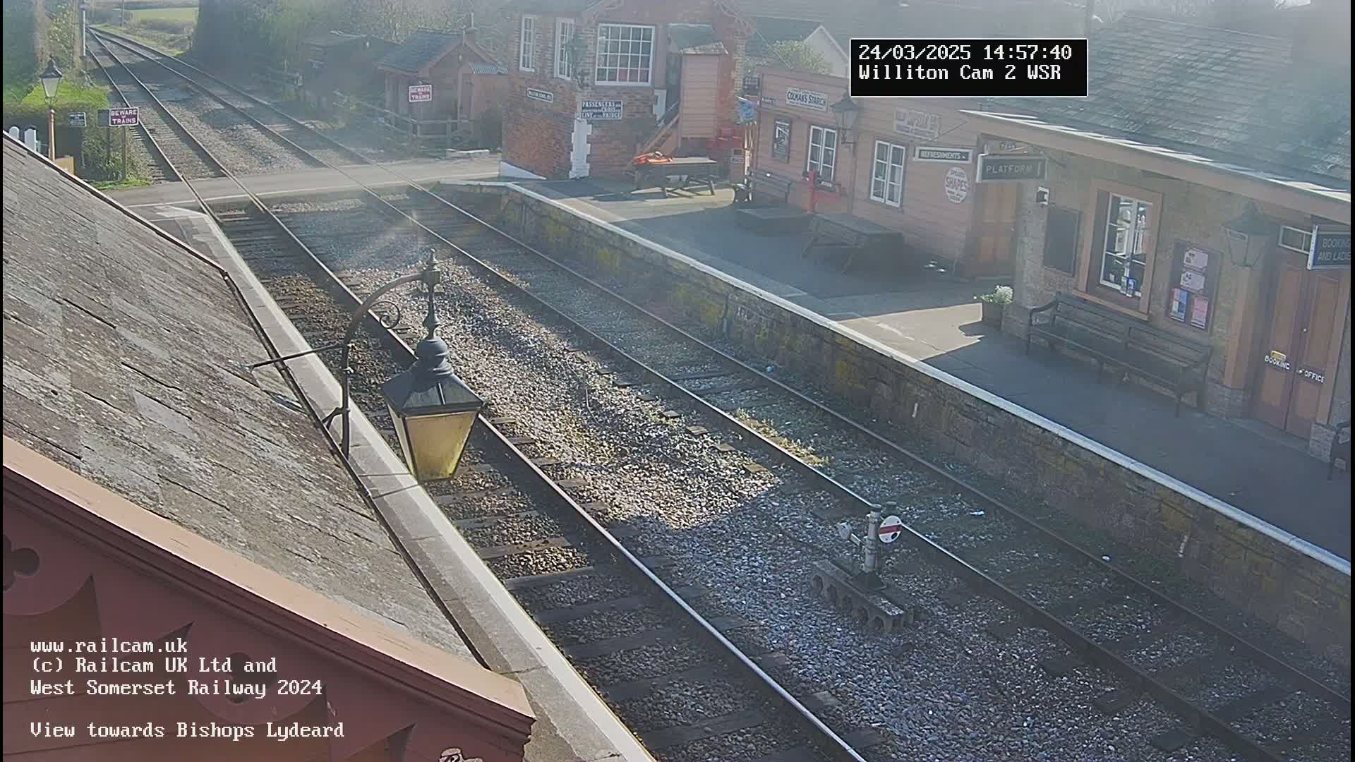 A sunny view of a train station with multiple tracks and a small building, seen from an elevated vantage point.