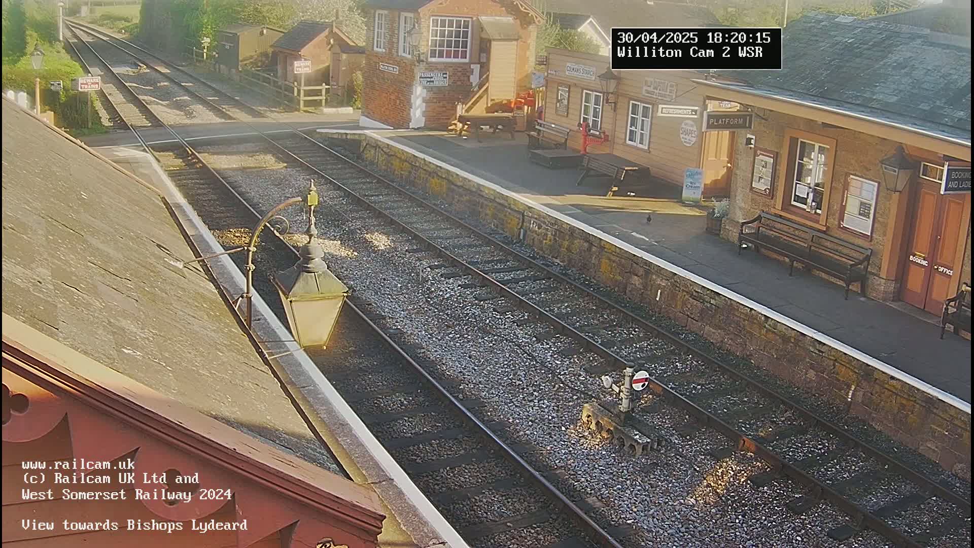 A sunny view of a train station with two parallel train tracks, a platform, and several buildings.