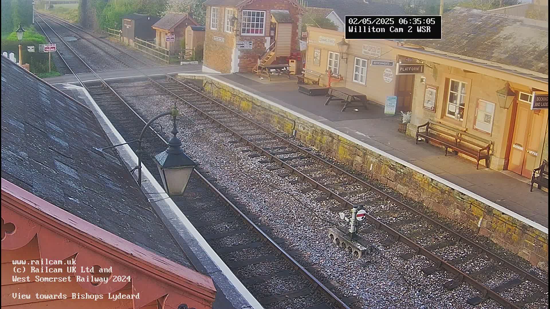 A high-angle view of a train station platform on a sunny morning, showing train tracks extending into the distance.