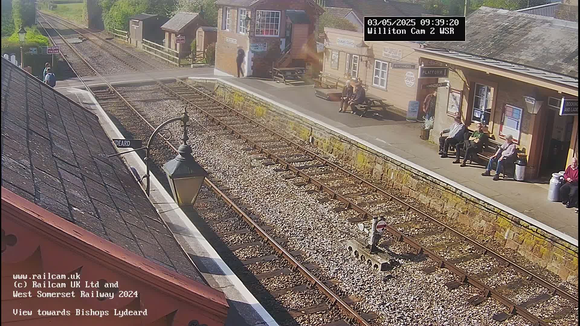A sunny day at a train station with several people sitting on benches and a couple standing near the tracks.