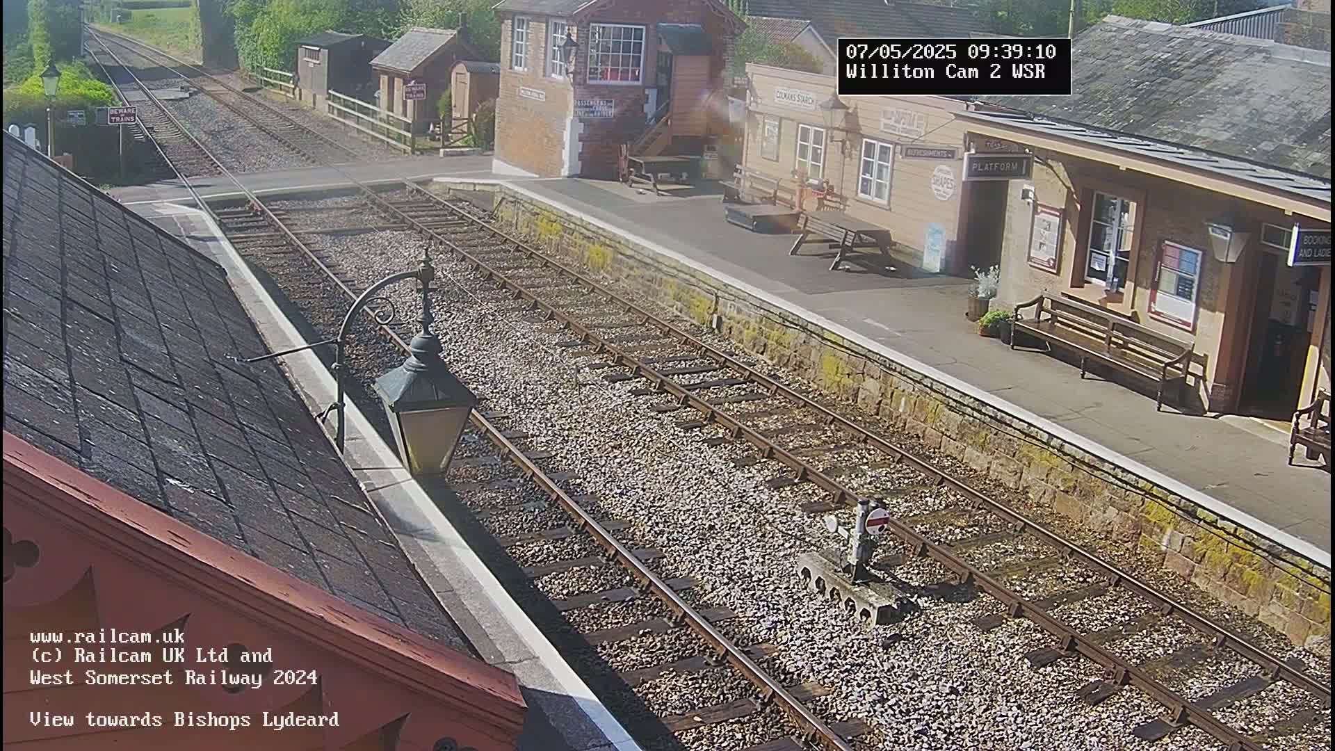 A sunny view of a train station platform with two sets of train tracks extending into the distance.