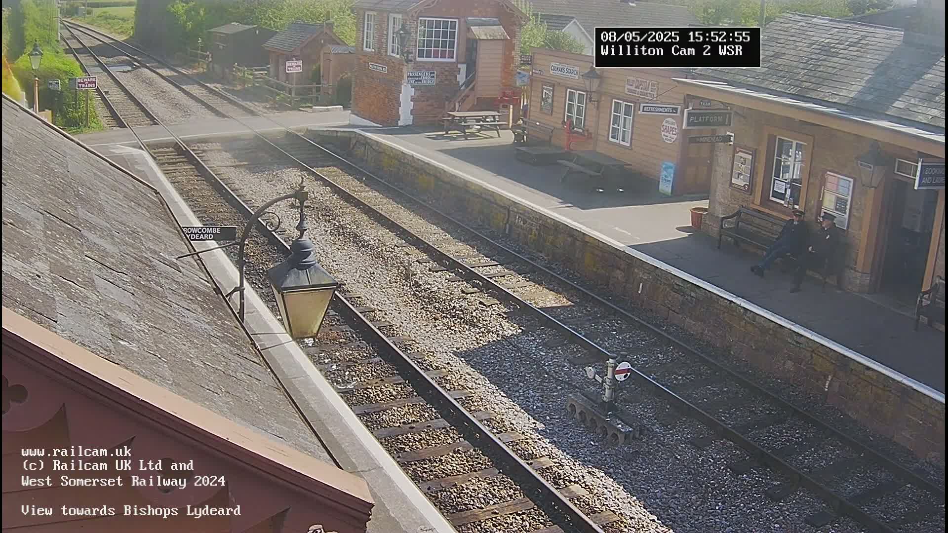 A sunny view of a train station platform with two men sitting on a bench, multiple train tracks extending into the distance, and station buildings visible.