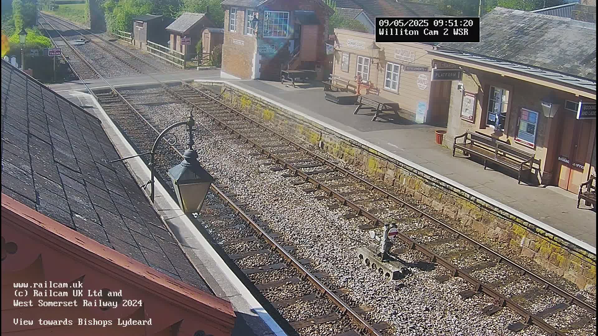 A sunny view from a train station platform shows railway tracks extending into the distance, with a station building and benches visible.