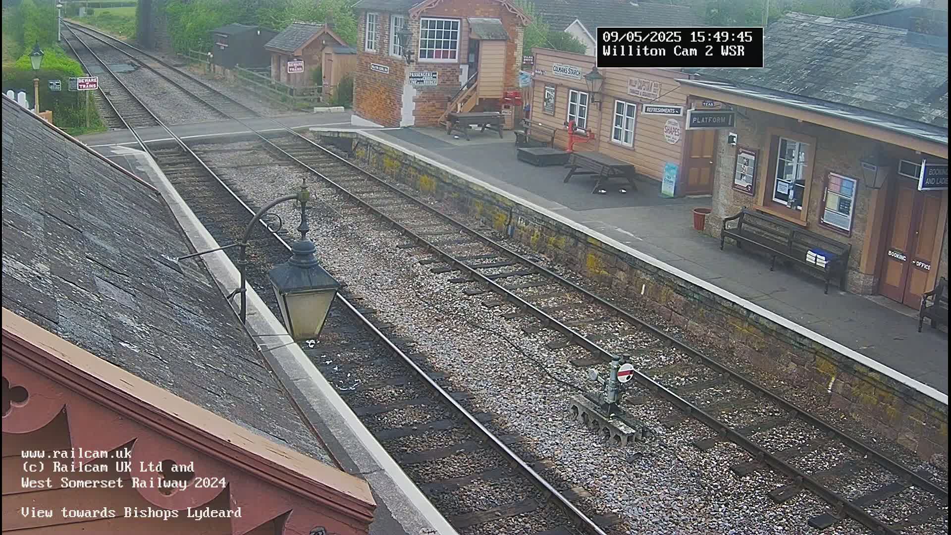 A high-angle view shows a railway station platform with train tracks extending into the distance under an overcast sky.