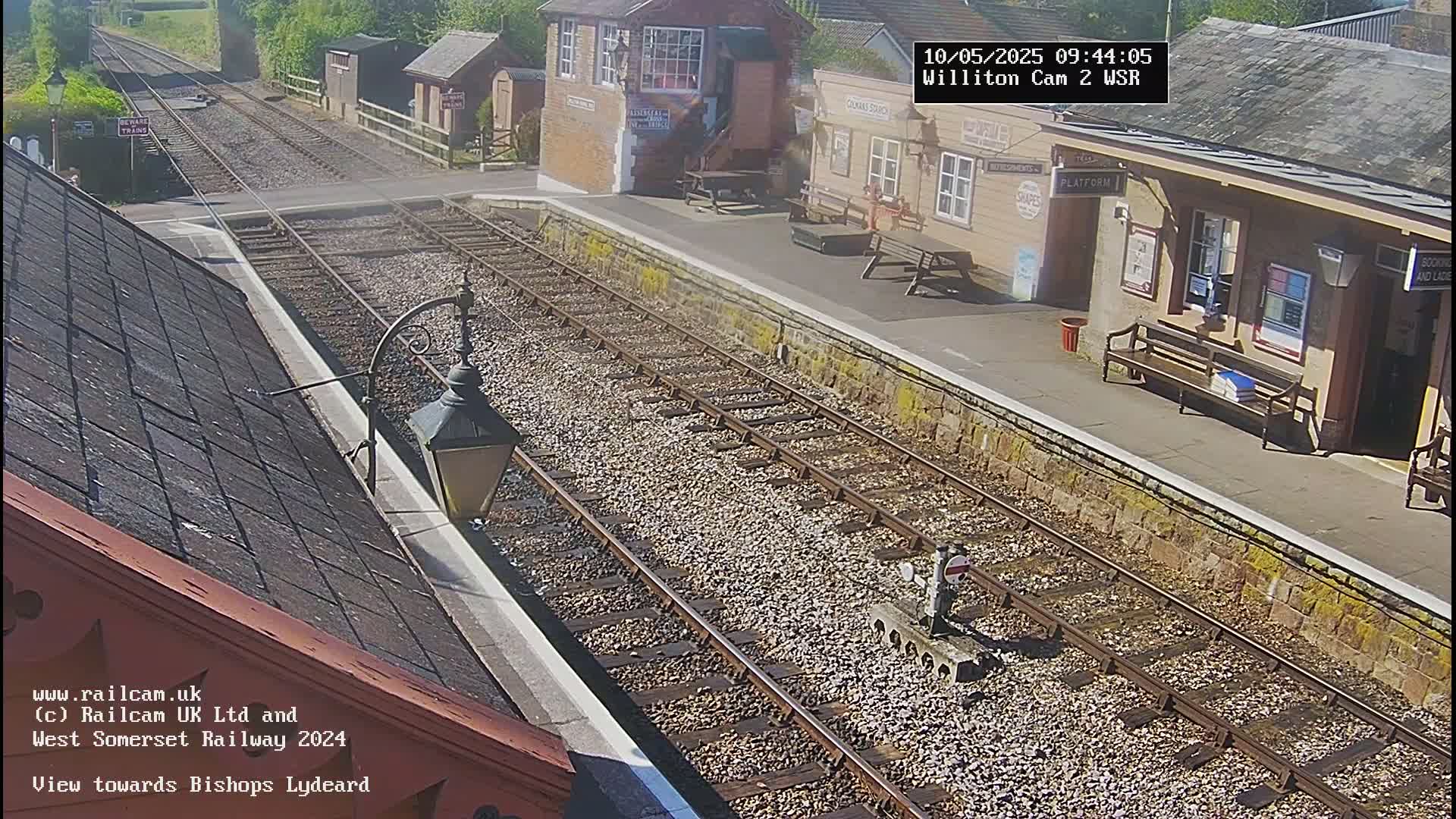 A sunny view of a train station platform with two tracks and several benches, seen from a high vantage point.