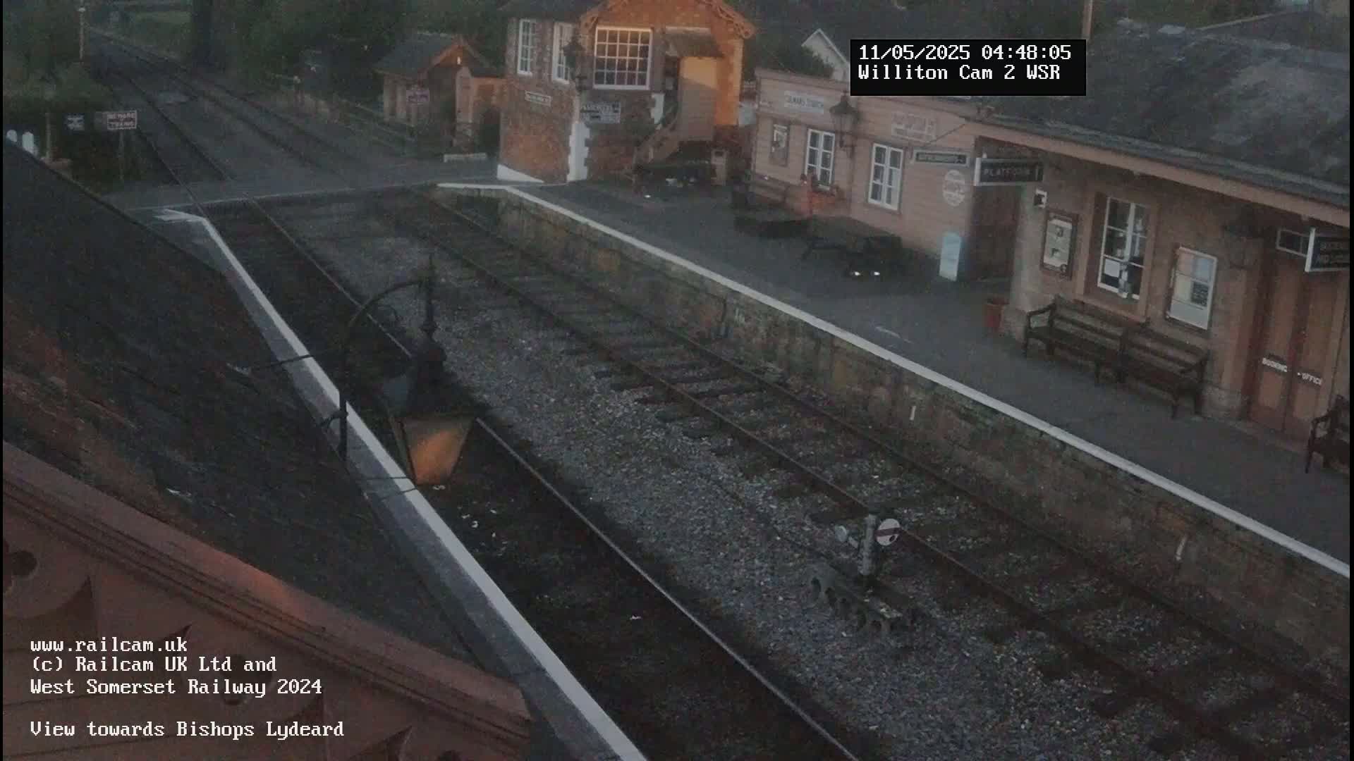 A train station platform with a few benches and buildings is visible on an overcast day, with train tracks extending into the distance.