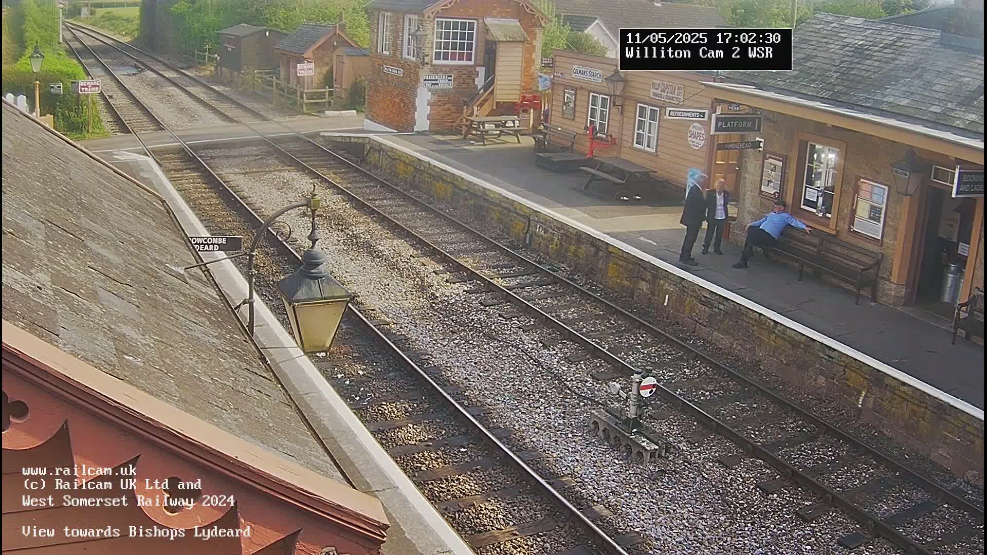 A sunny view of a train station with multiple tracks, a platform with several people, and buildings in the background.