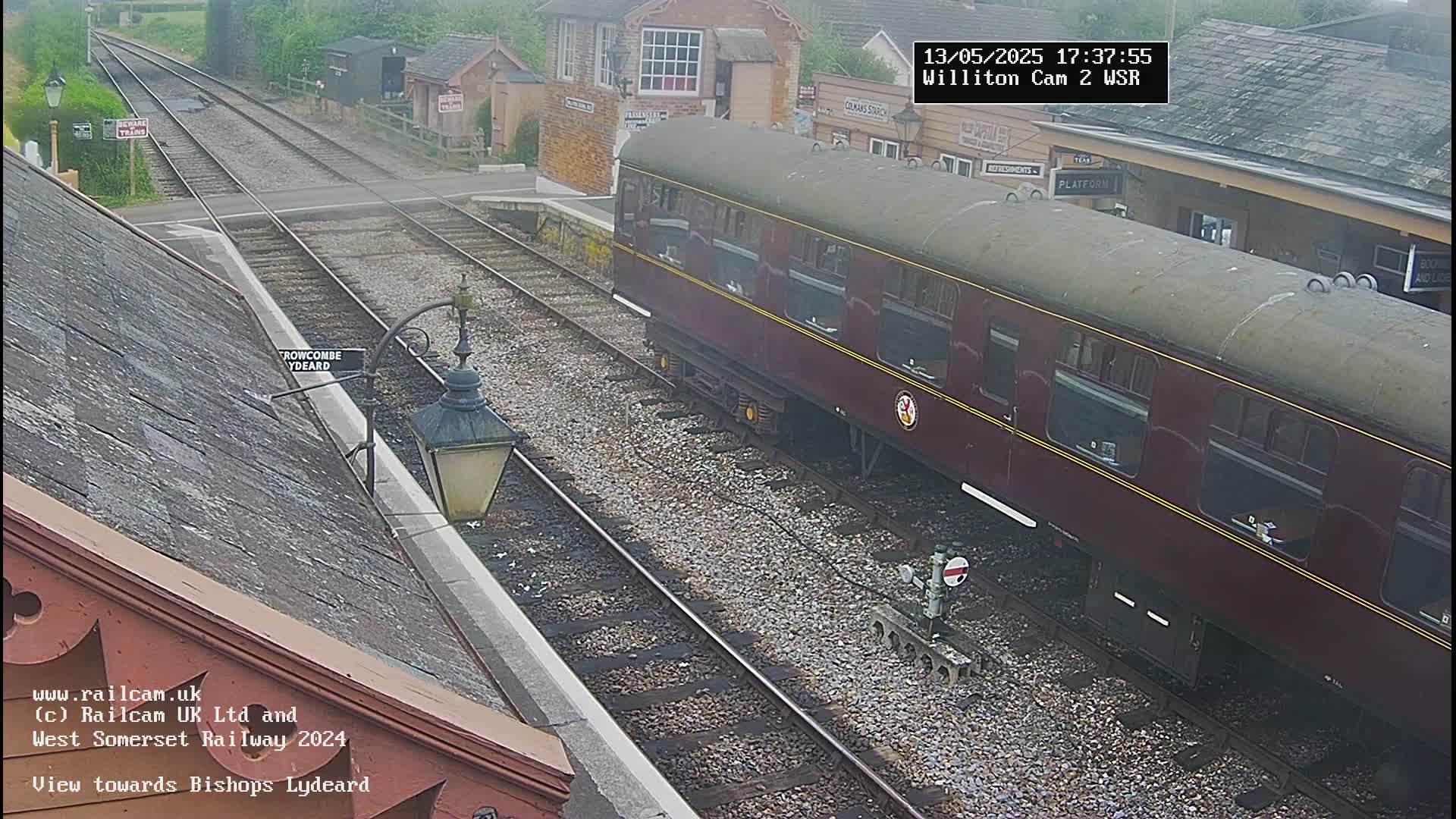 A maroon train car sits at a railway station on a cloudy day, next to a track splitting off in two directions.