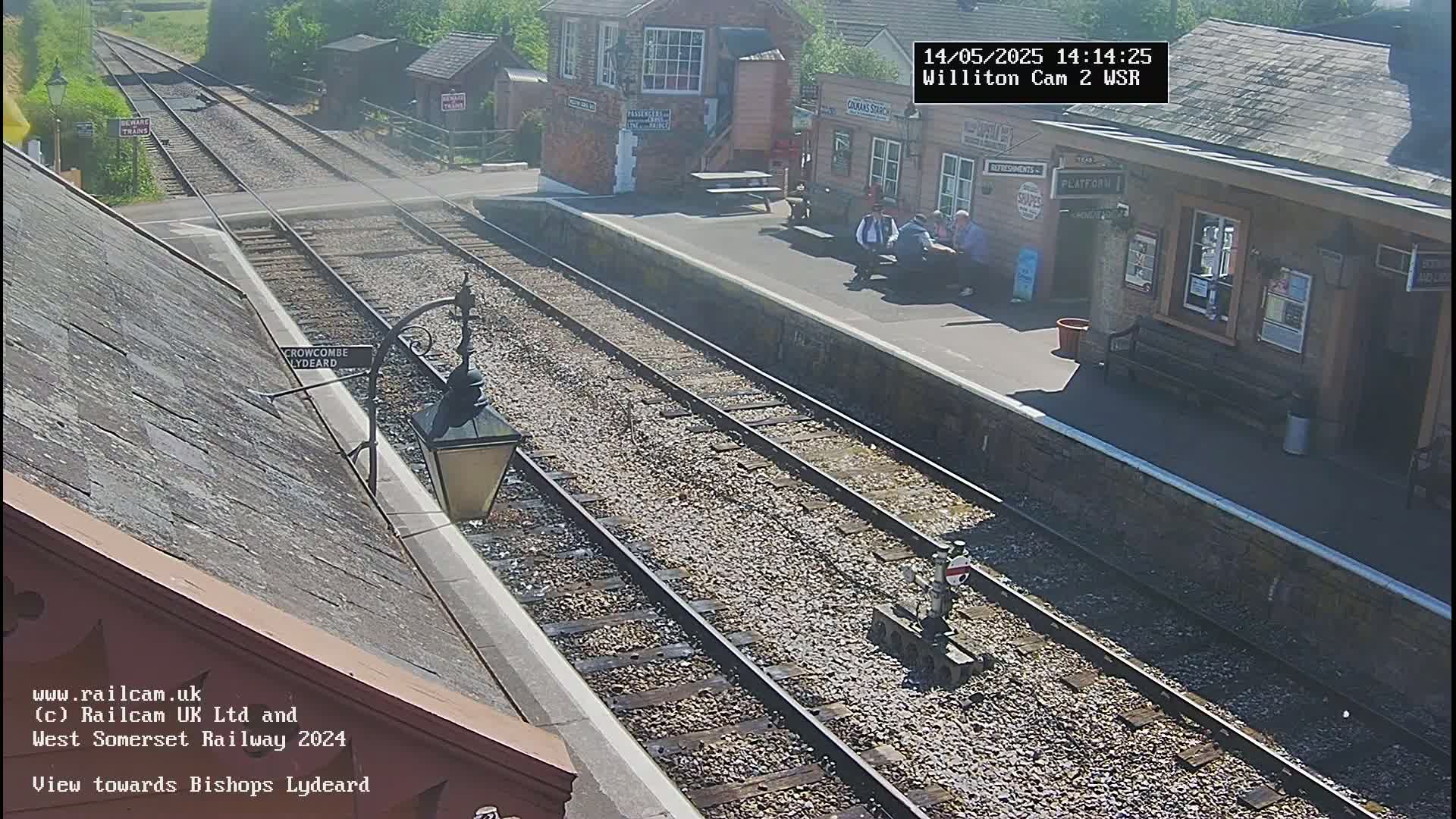 A sunny view of a train station platform with two sets of train tracks, a few people sitting on a bench, and several buildings.