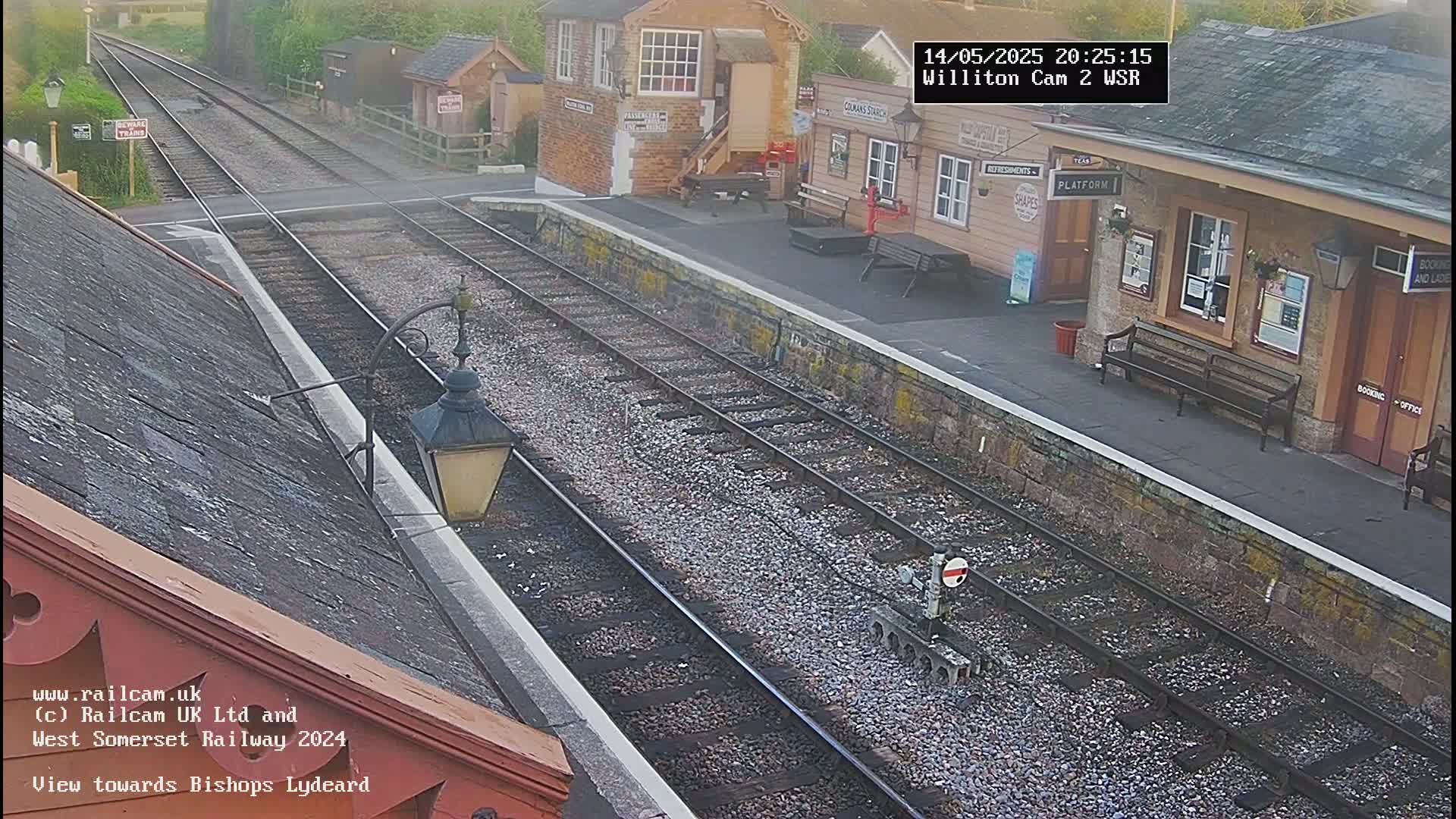 A train station platform and tracks are viewed from above on an overcast day, with a small train visible in the distance.