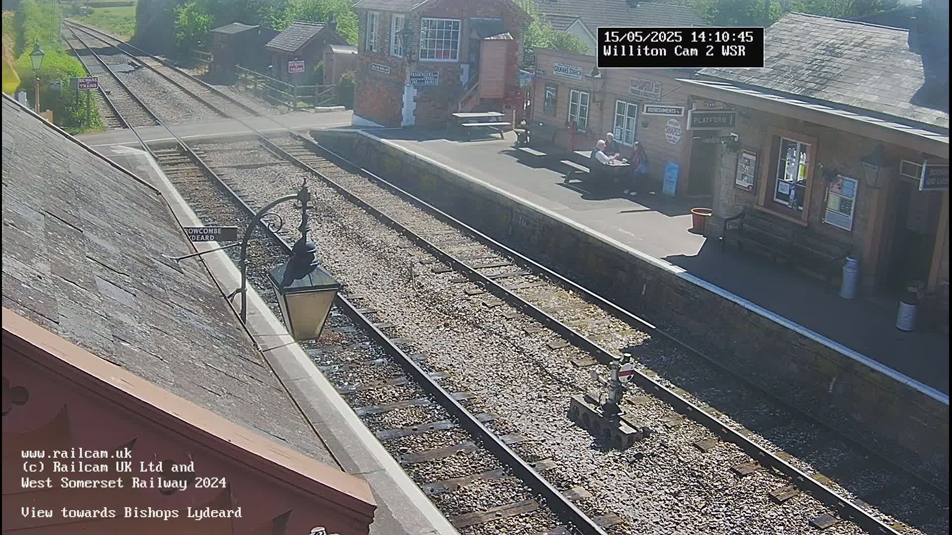 A sunny view of a train station platform with two parallel train tracks extending into the distance, and a few people sitting on a bench.