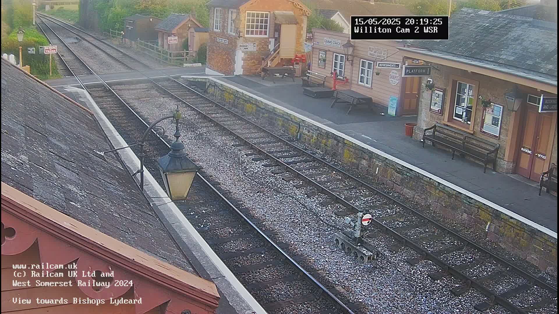 A train station platform with two sets of train tracks, a small railway signal, and several buildings under an overcast sky.
