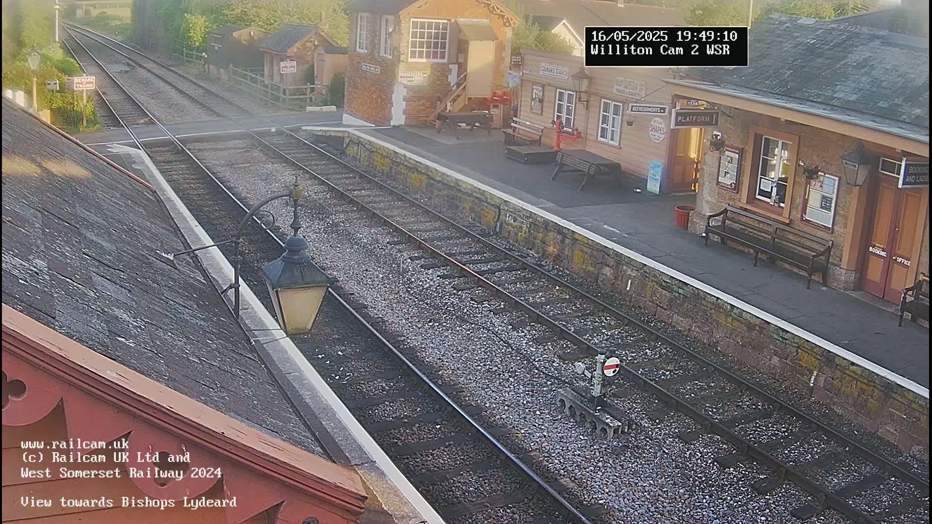 A high-angle view of a train station platform with multiple train tracks, under slightly hazy daylight conditions.