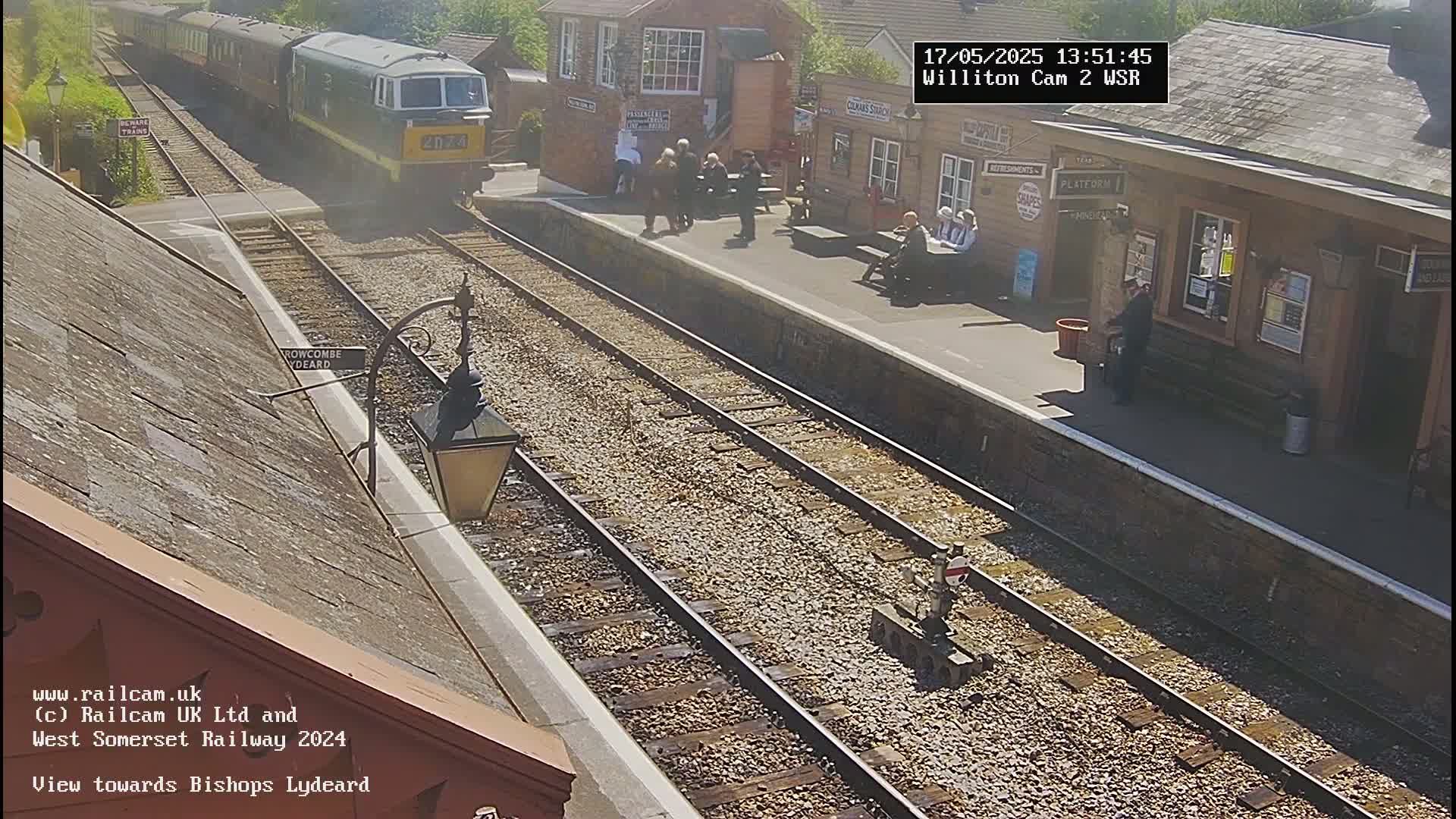 A train is departing a sunny railway station platform where several people are standing and sitting.