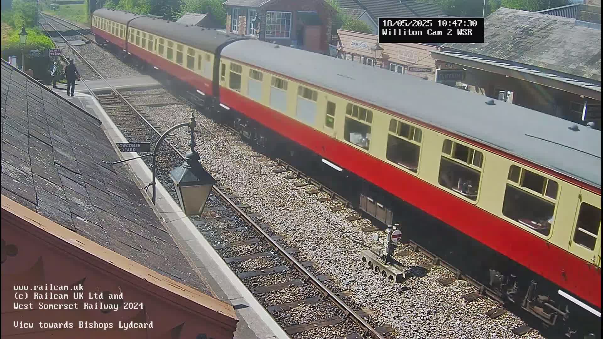 A red and cream passenger train is slowly moving through a sunny train station.