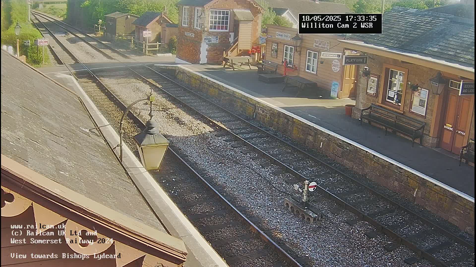 A sunny view of a train station platform with two sets of train tracks extending into the distance.