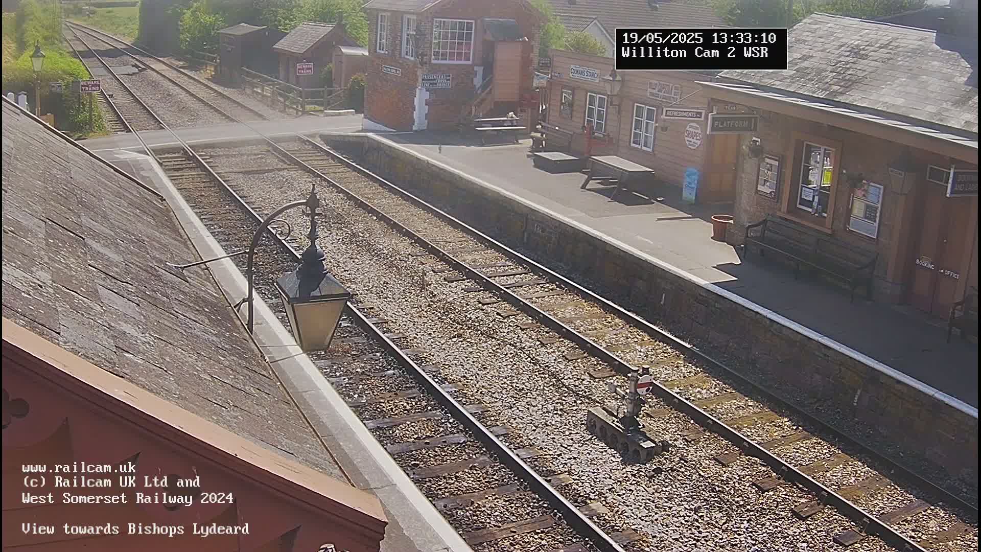 A sunny view of a railway station platform with two sets of train tracks extending into the distance.