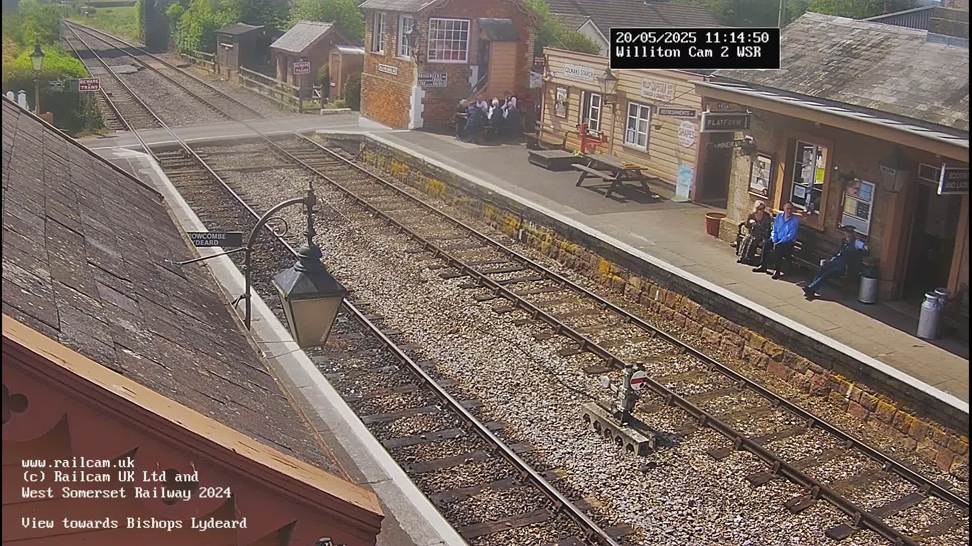 A sunny day at a train station shows several people sitting on benches near a platform, with train tracks extending into the distance.
